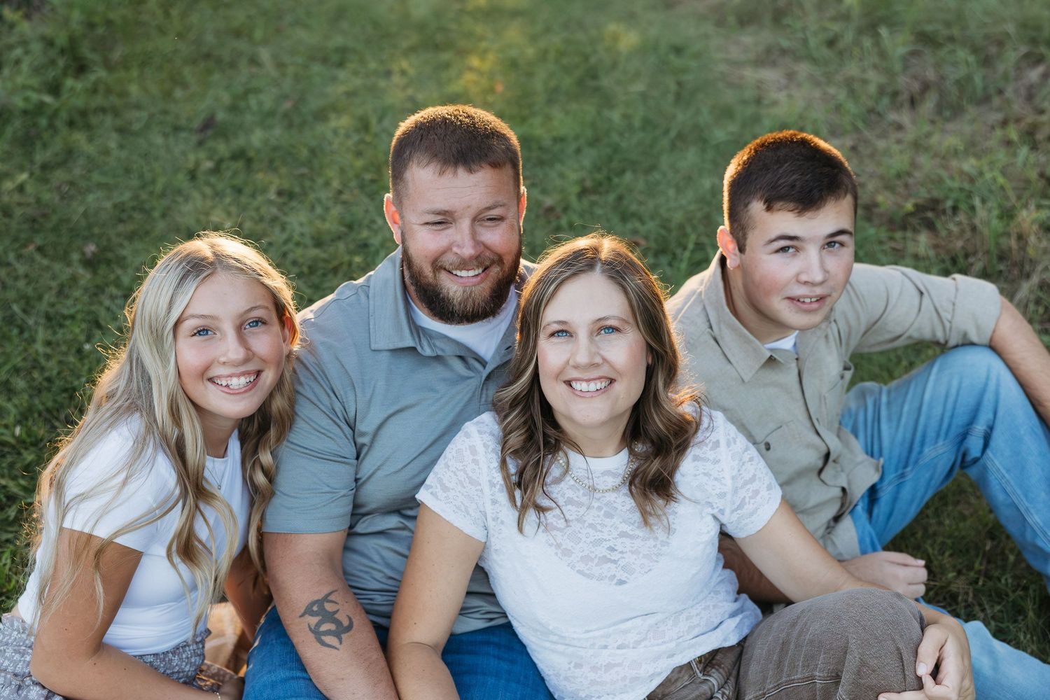 A smiling family of four poses together on a grassy hillside during a warm outdoor photo session.