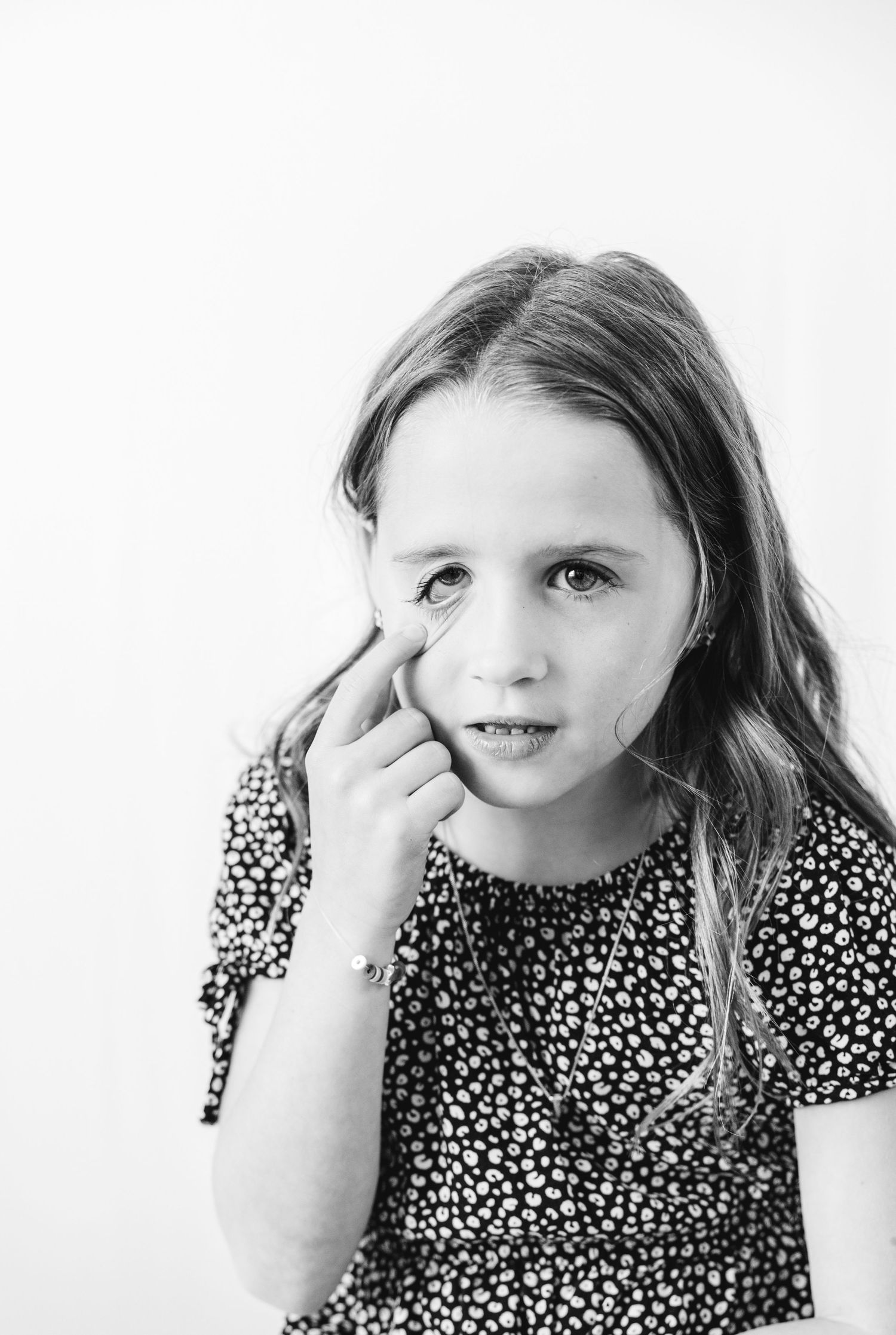 Black and white portrait of a young girl with long hair touching her face, wearing a floral dress.