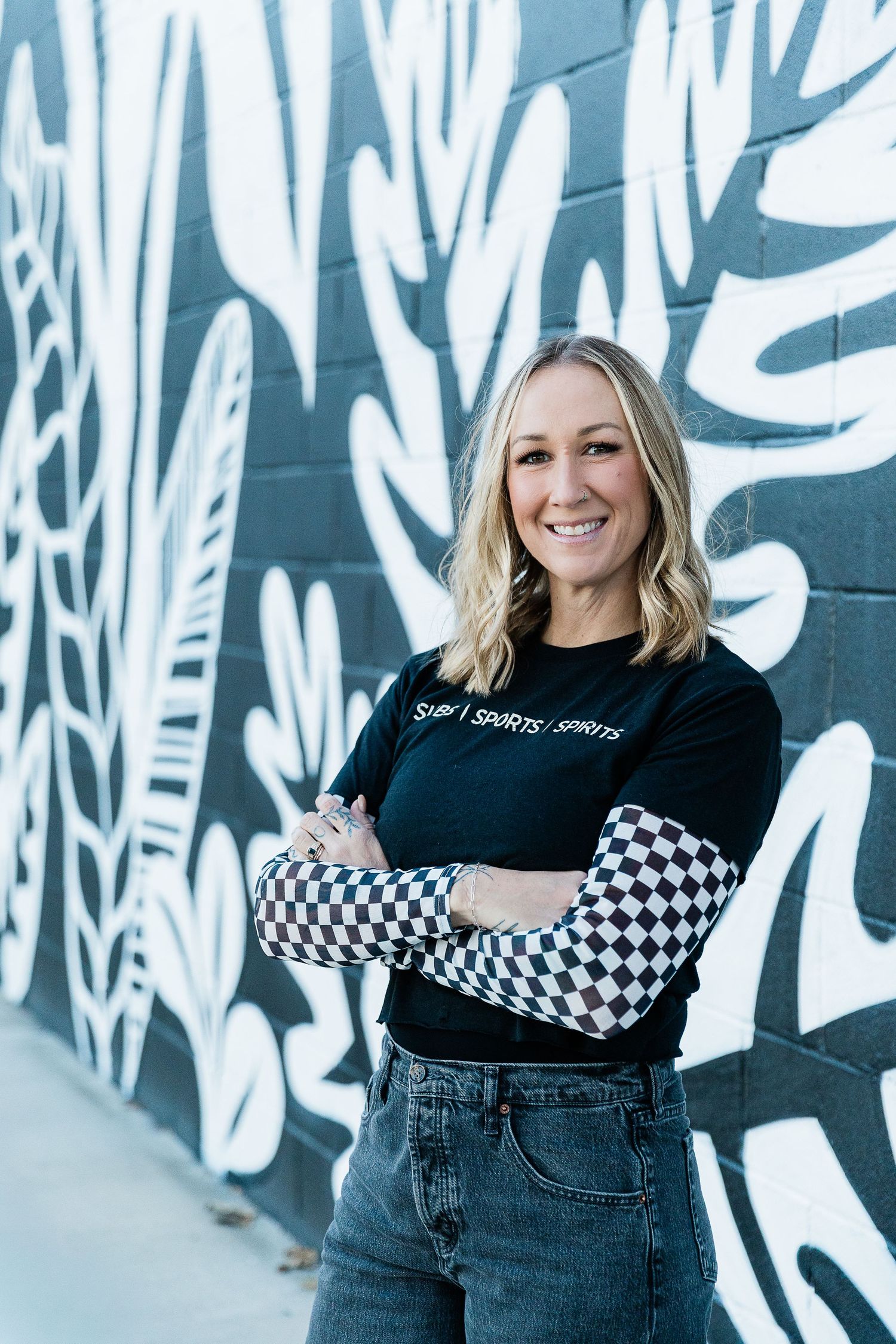 Smiling blonde woman in black Subs Sports Spirits t-shirt with checkered sleeves, arms crossed, posing in urban alley. Katie Chamberlain