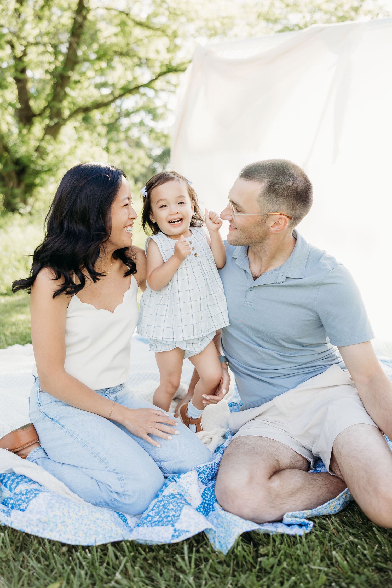 Happy family with toddler daughter enjoying a sunny outdoor picnic on a blue quilt blanket in a lush green park.