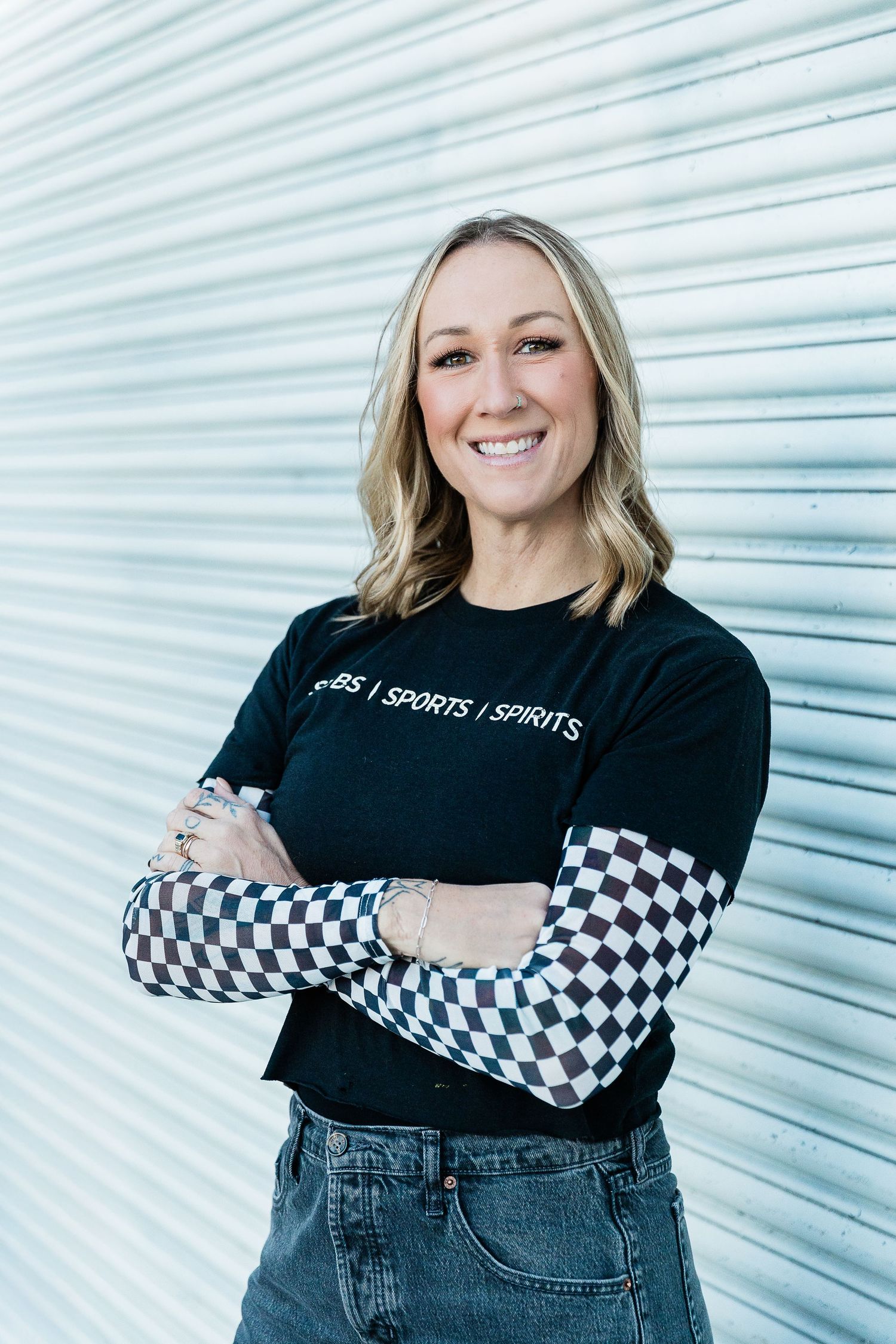 Smiling blonde woman in black Subs Sports Spirits t-shirt with checkered sleeves, arms crossed, posing in urban alley. Katie Chamberlain