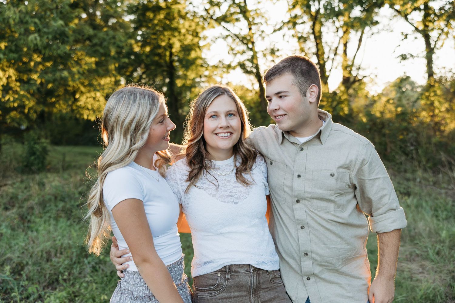 Three smiling friends pose together outdoors in golden hour light, surrounded by lush green trees.