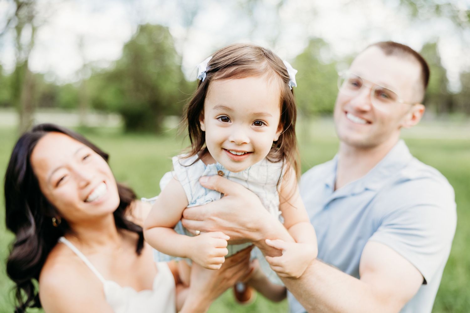 Happy family with parents holding smiling toddler girl outdoors in green park setting.