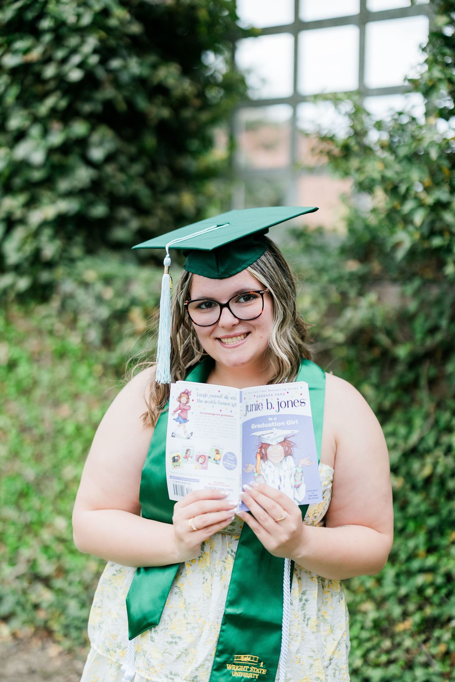 Smiling graduate in green cap holds a childrens book outdoors in front of green foliage. Photographed by Katie Chamberlain Photography