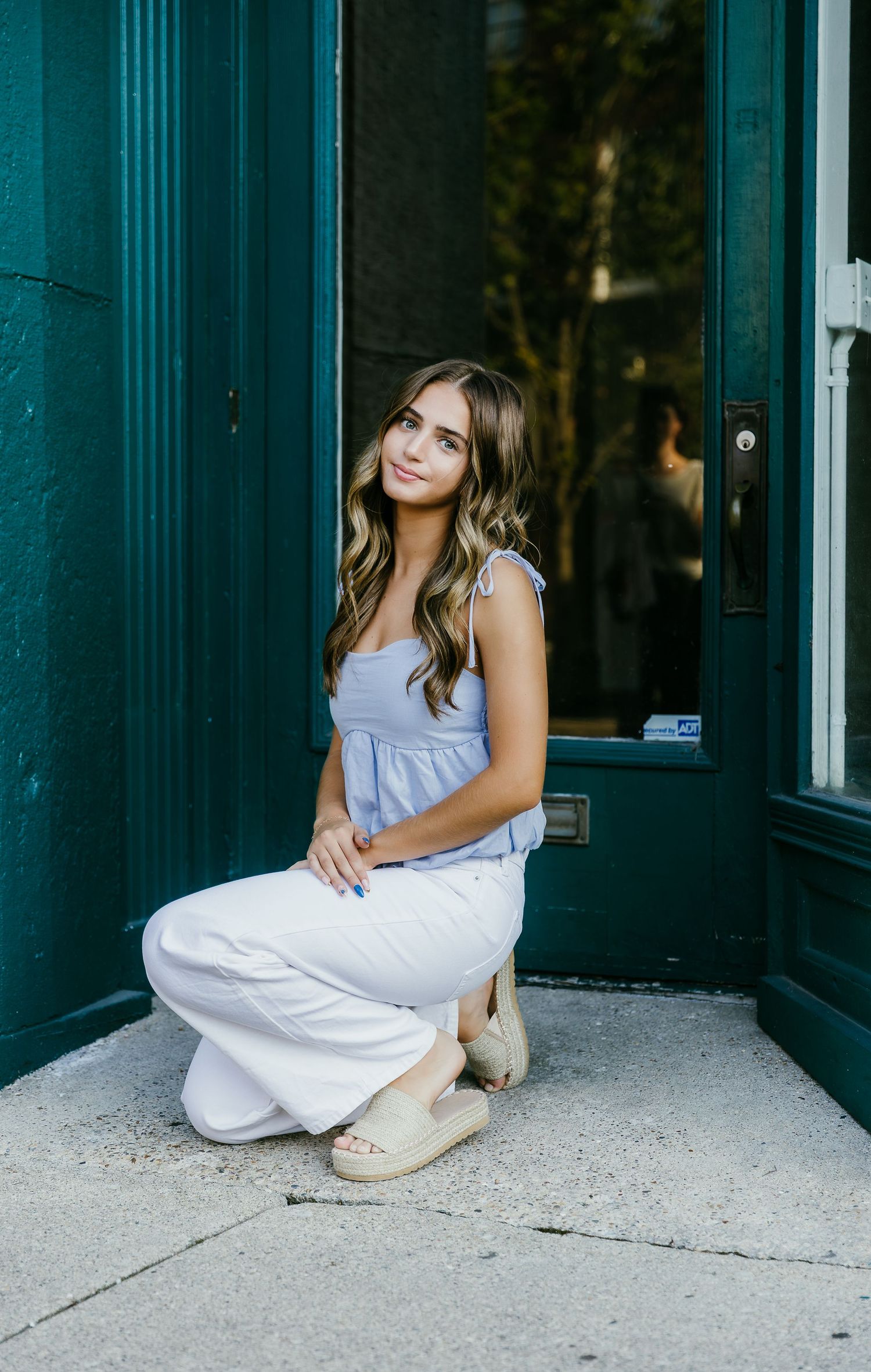 Young woman in blue top and white wide-leg pants posing by a teal door on a city sidewalk.
