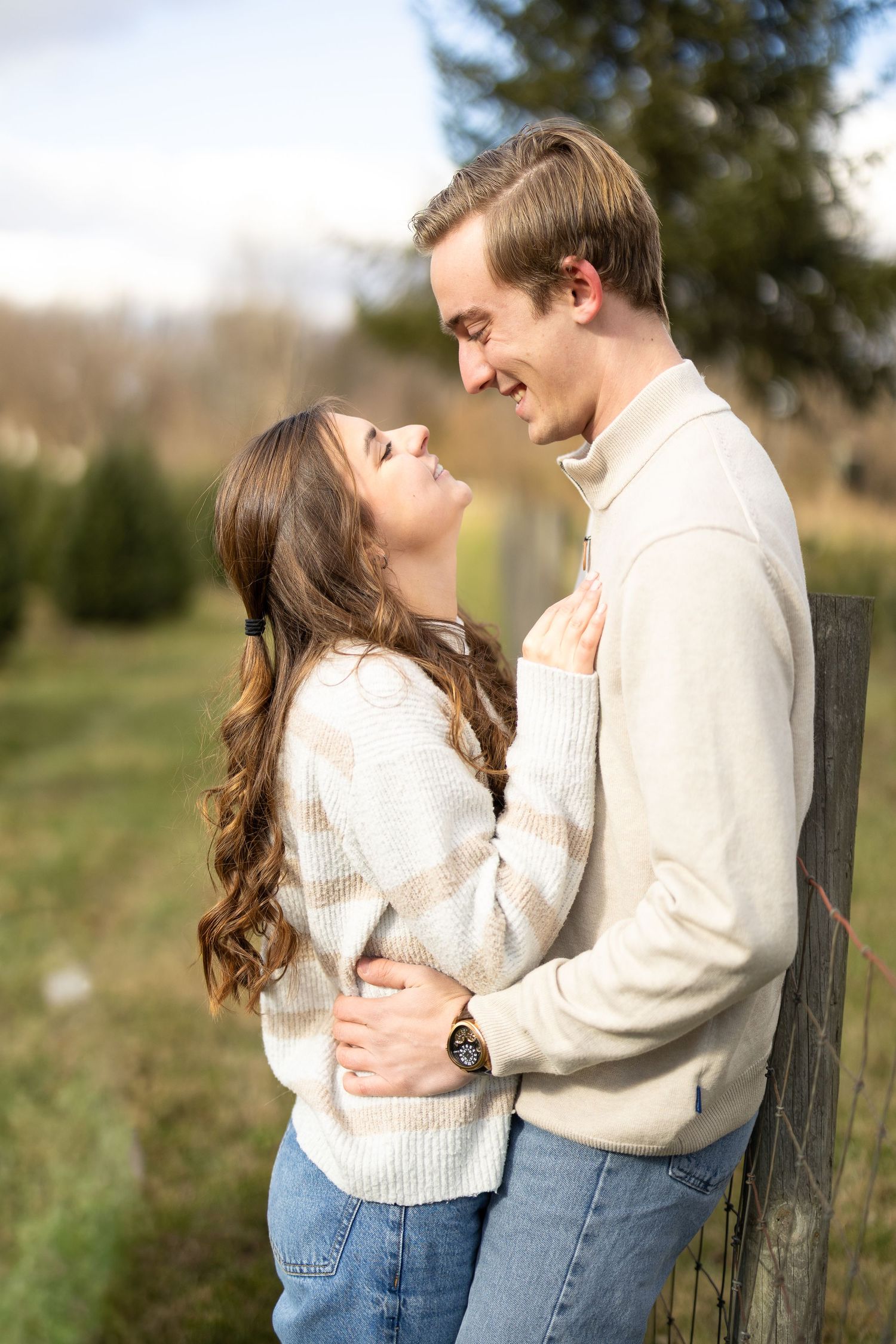 A smiling couple in matching cream sweaters and jeans embrace outdoors near a wooden fence, evergreen trees behind them. Katie Chamberlain