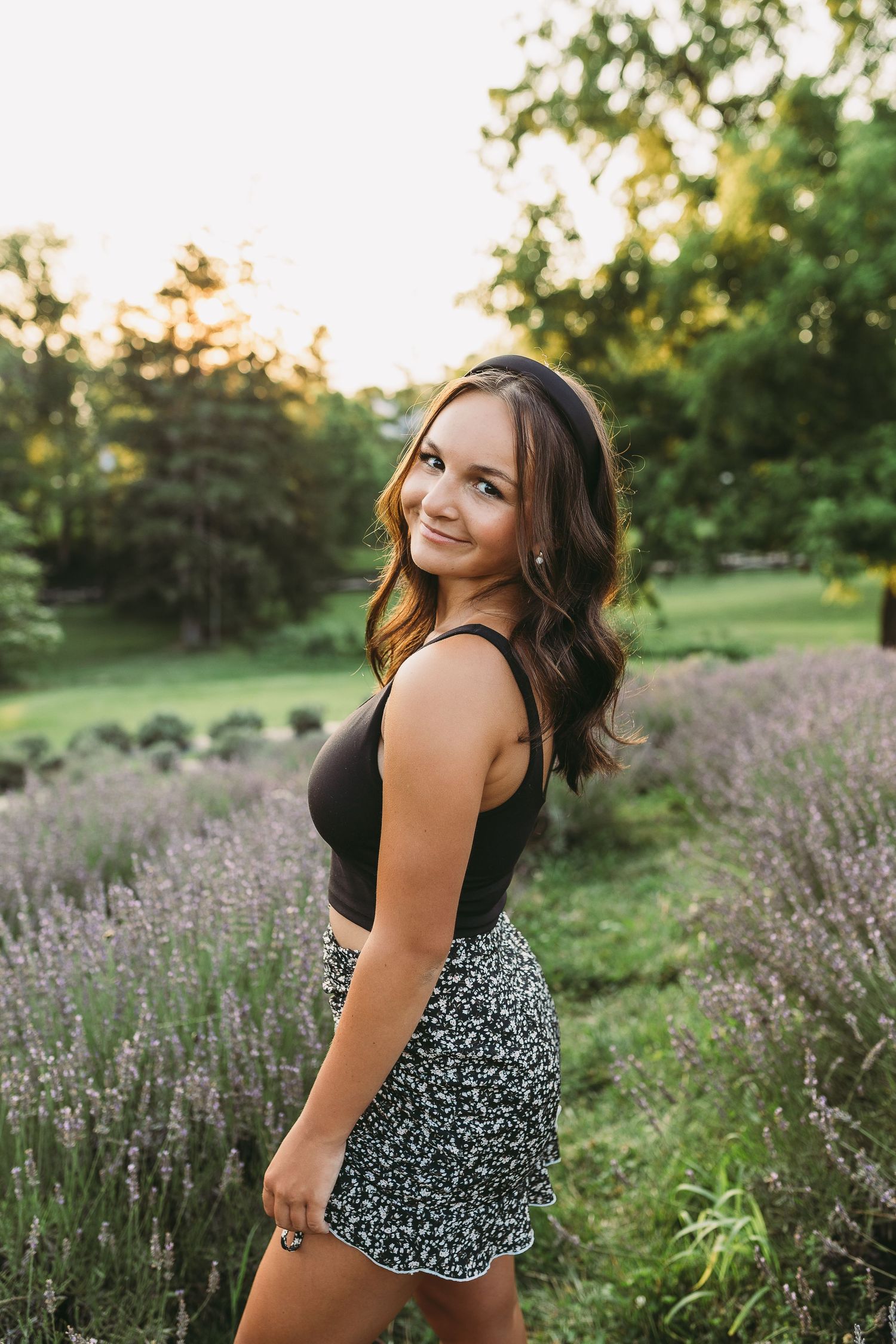 Young woman in black top and floral skirt smiling in a lavender field at golden hour. Katie Chamberlain Photography