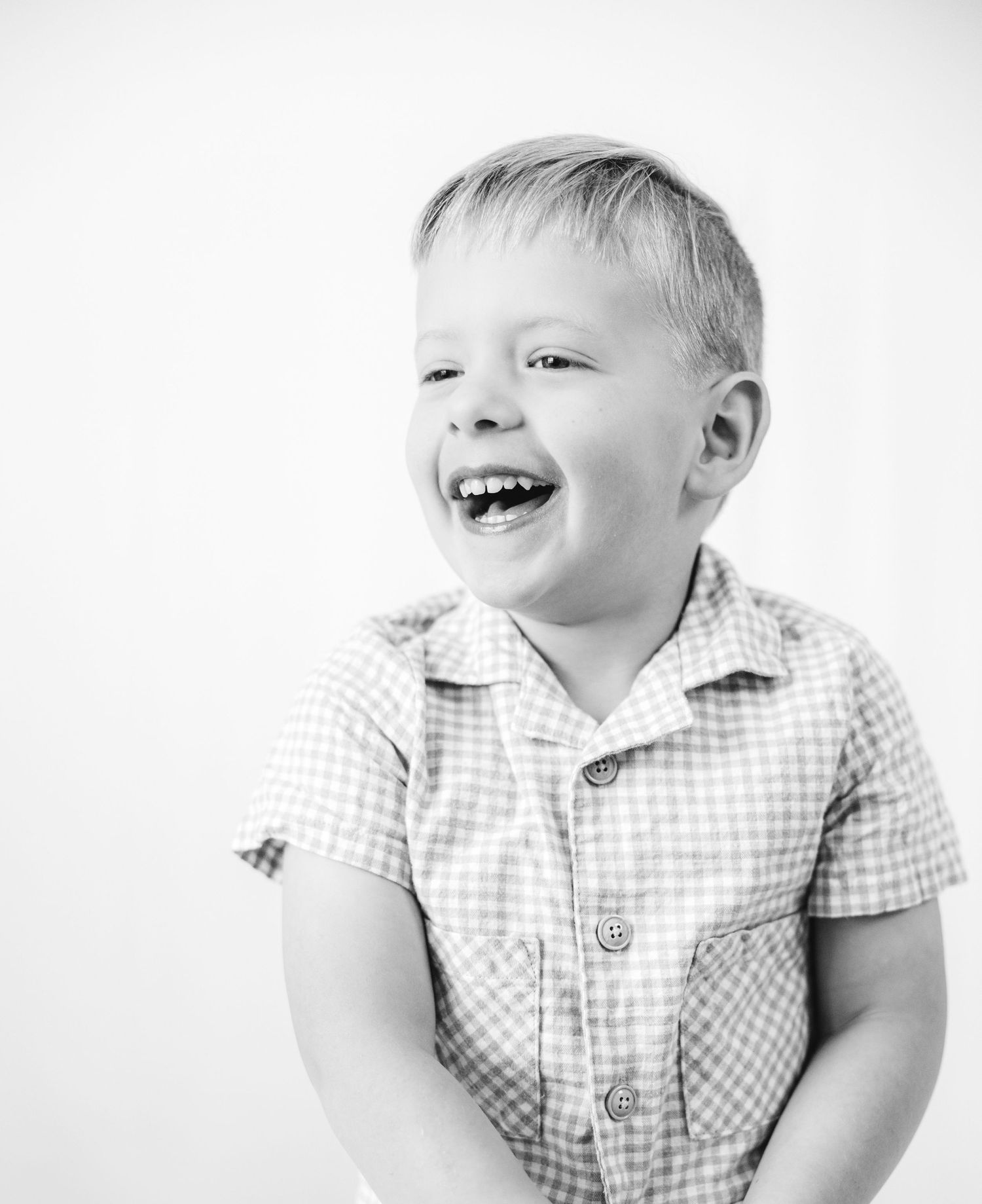 Black and white portrait of a laughing blonde toddler boy wearing a checkered shirt against white background.