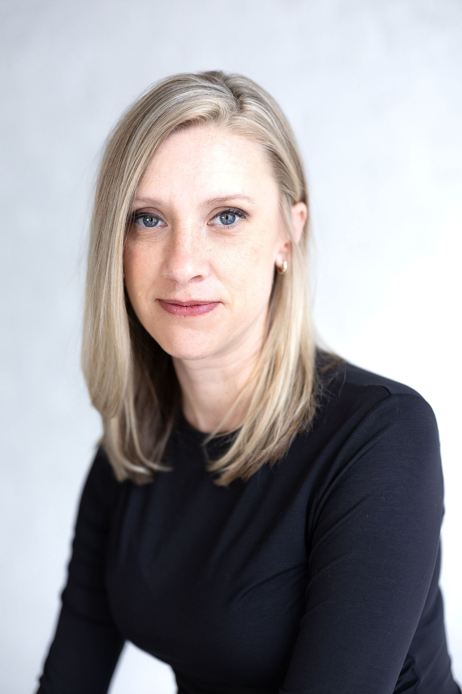 Professional headshot of a blonde woman in a black top against a light gray background. Katie Chamberlain Photography