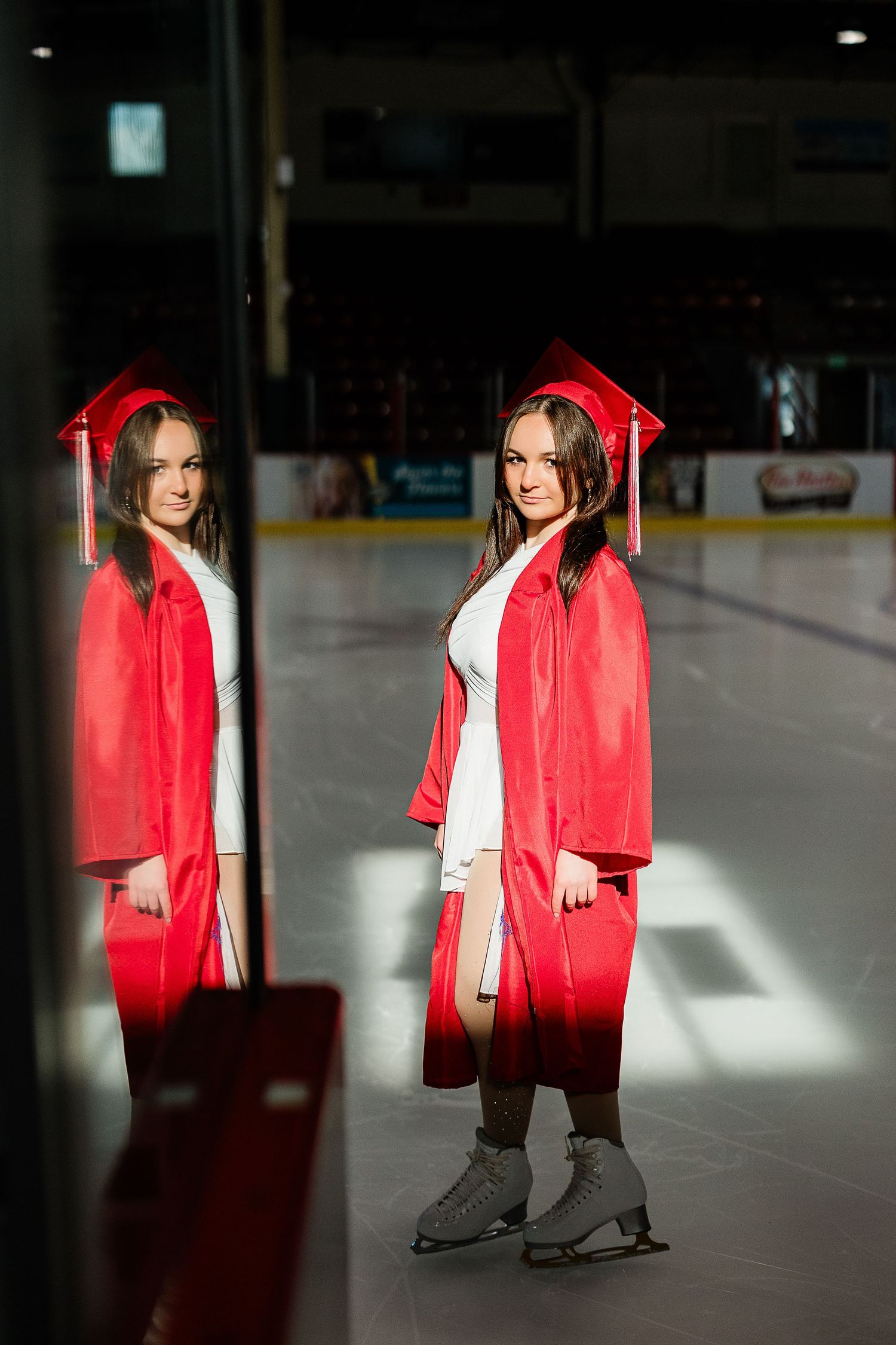 Graduate in red cap and gown poses on ice rink in skates, reflected in rinkside glass. Katie Chamberlain Photography