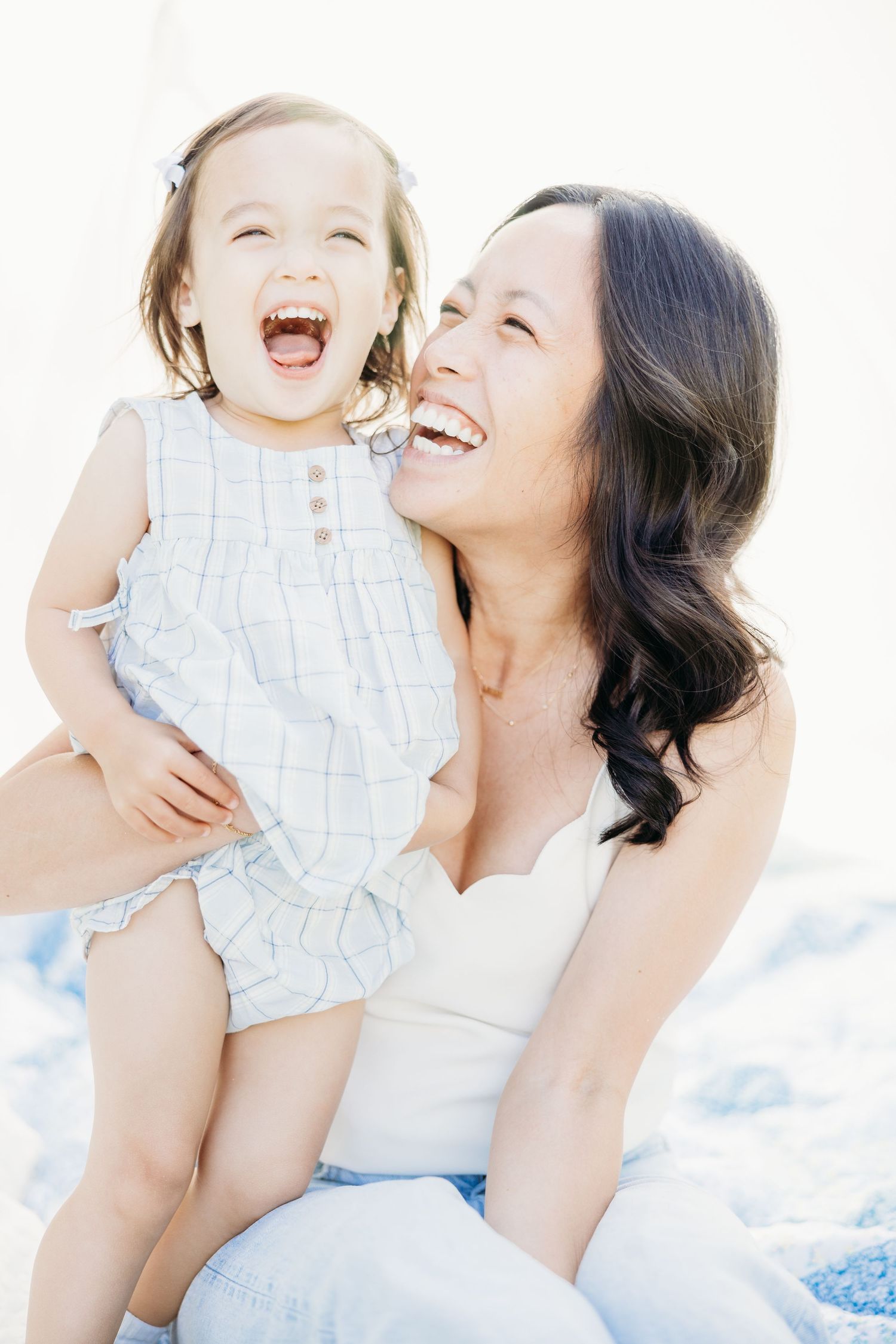 A joyful mother holds her laughing toddler daughter outdoors in bright natural light, both smiling radiantly.