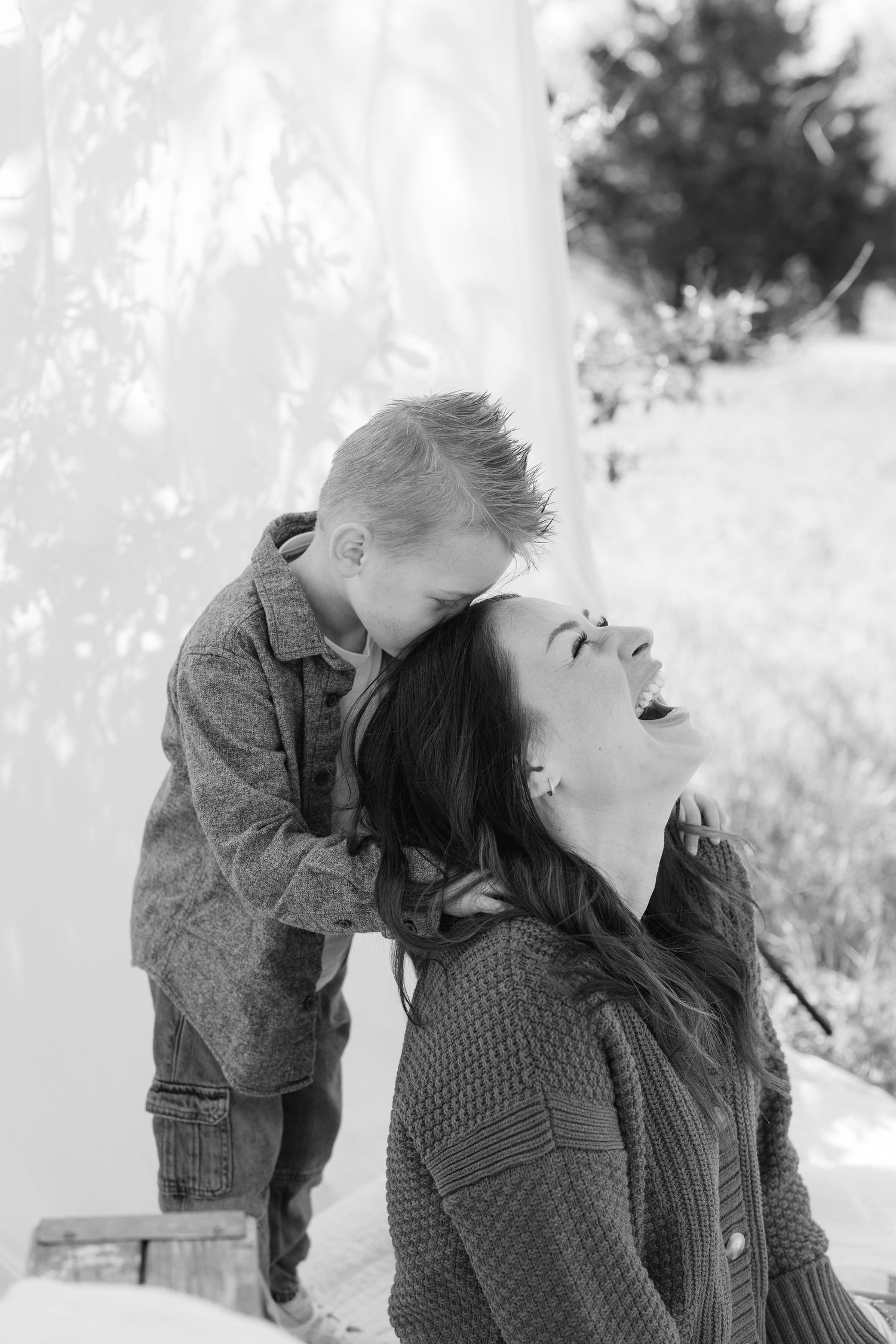 Black and white photo of a laughing mother and young son sharing a joyful moment outdoors near a white fabric backdrop.