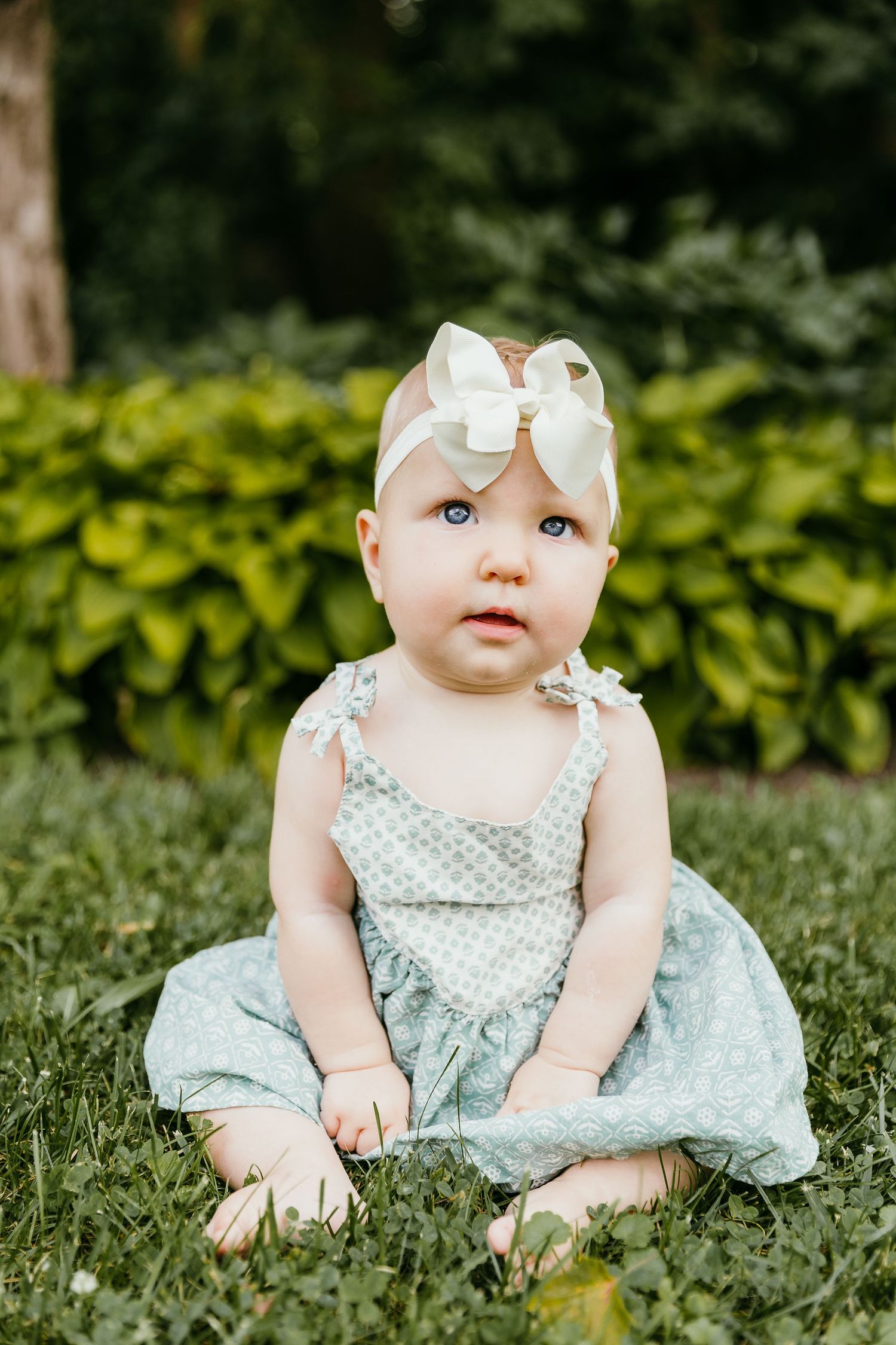 A cute baby girl in a light blue polka dot dress and white bow headband sits on green grass outdoors. Photographed by Katie Chamberlain