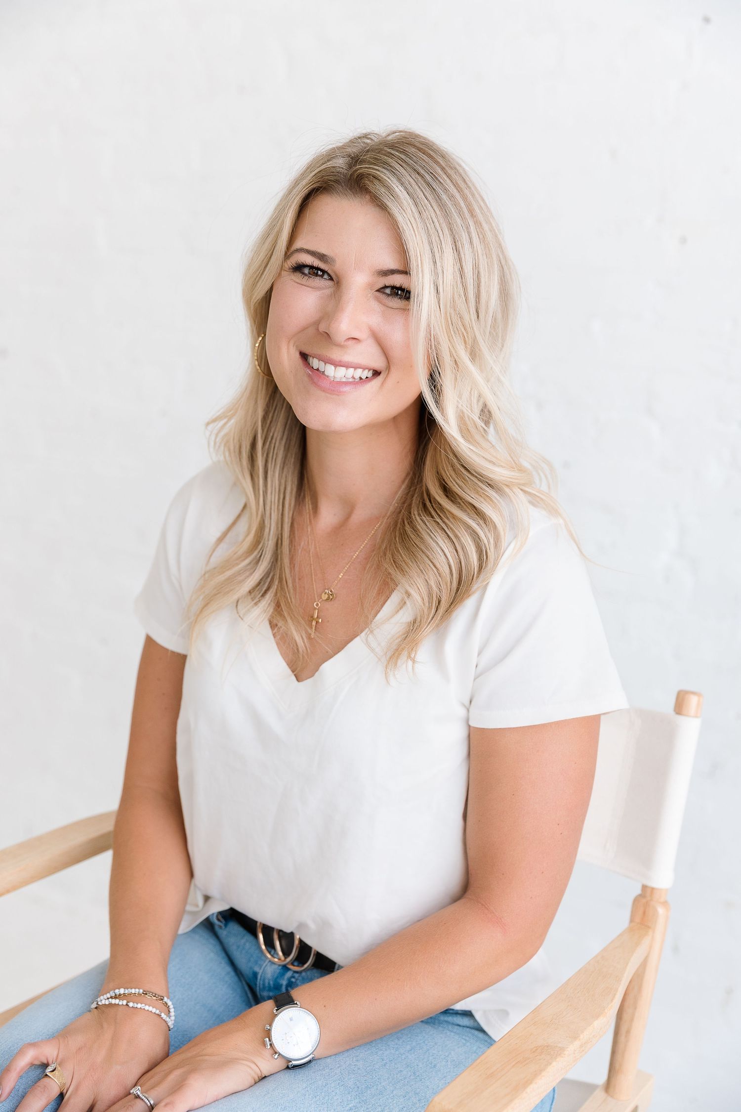 Smiling blonde woman in white v-neck tee and jeans sitting in a wooden chair against a white wall. Photo by Katie Chamberlain Photography