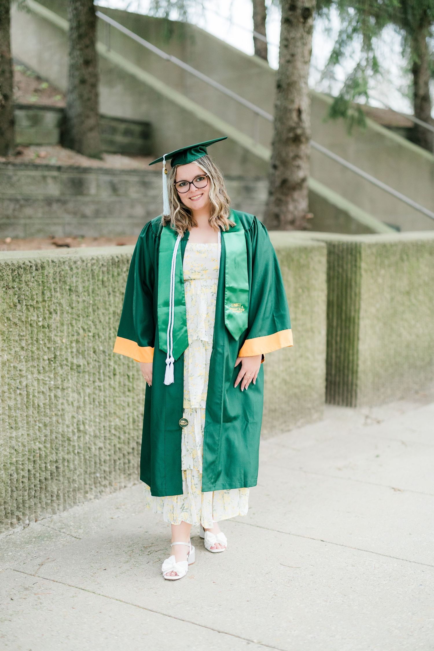 Smiling woman in green graduation gown and cap poses outdoors against a moss-covered wall. Photographed by Katie Chamberlain Photography