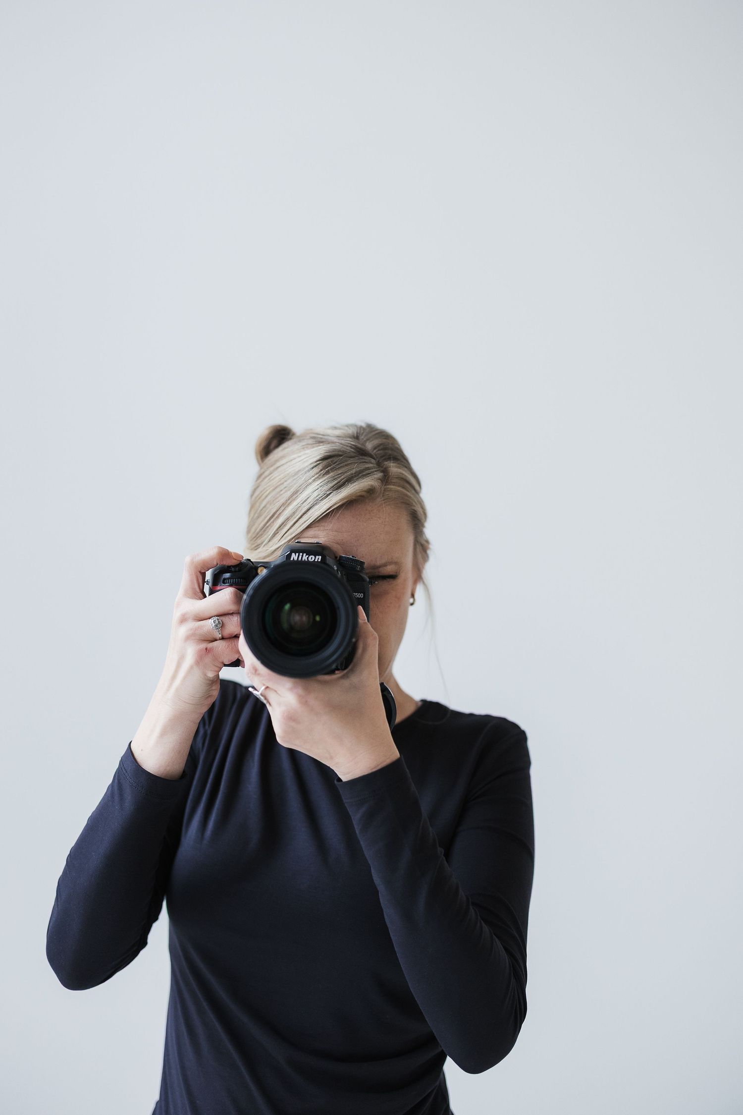 Blonde woman in black top holding a DSLR camera up to her eye against a white background. Katie Chamberlain Photography