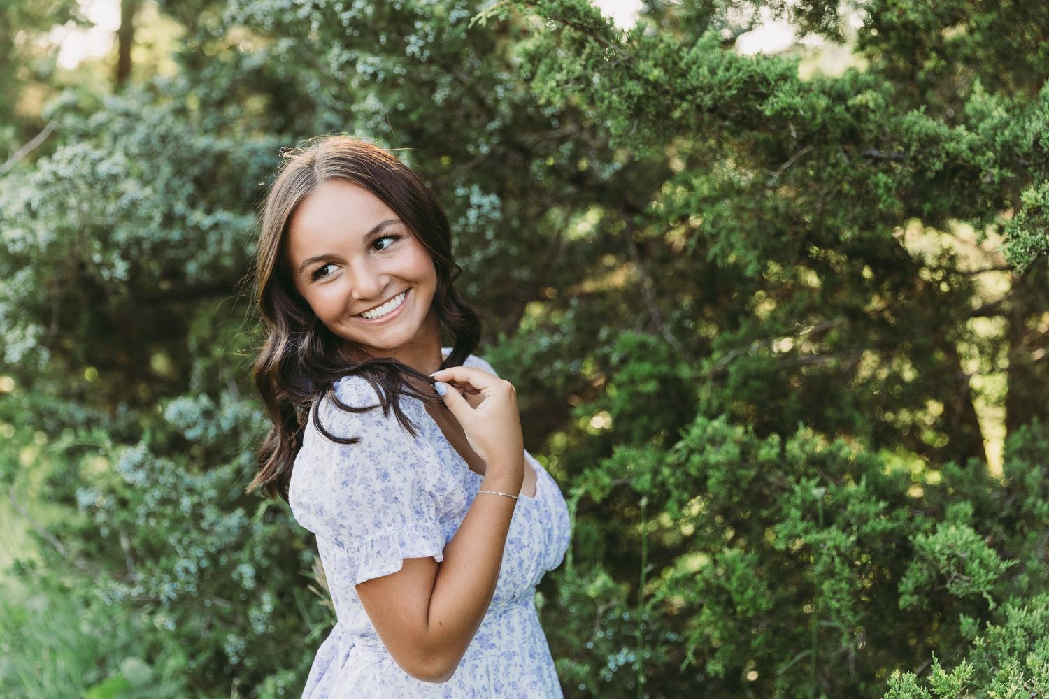 Smiling young woman in a white floral dress posing outdoors among lush green trees on a sunny day. Katie Chamberlain Photography