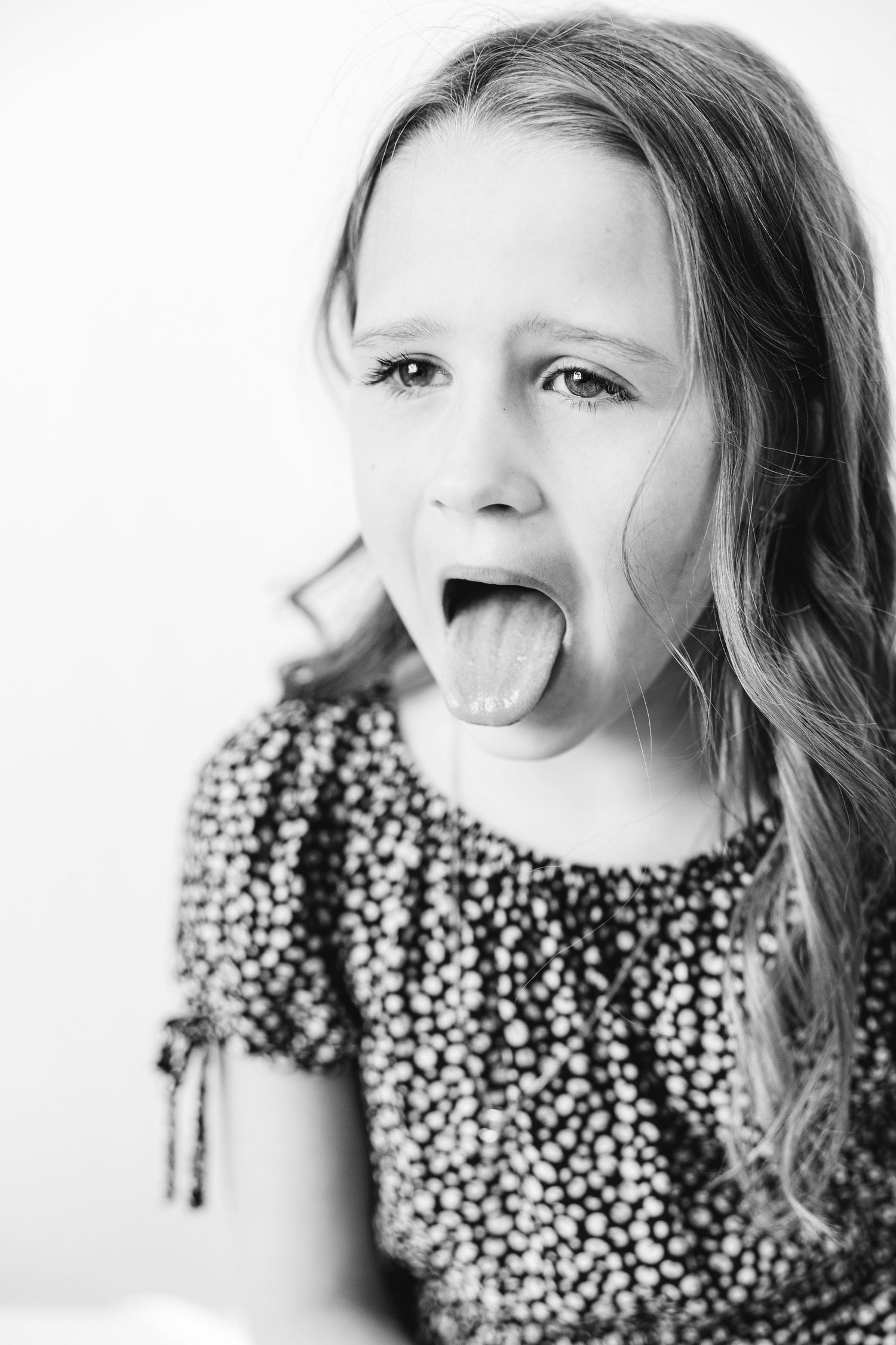 Black and white portrait of a young girl with long hair sticking out her tongue, wearing a polka dot dress.