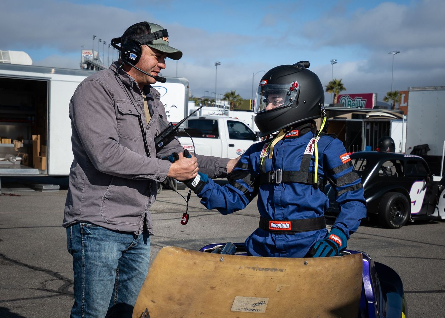 Racing coach with headset giving instructions to a young helmeted driver in a junior race car.