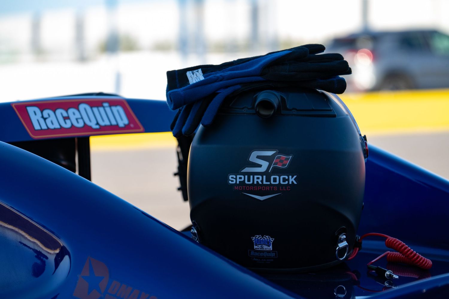 RaceQuip Spurlock branded racing helmet with gloves resting on a blue race car cockpit.