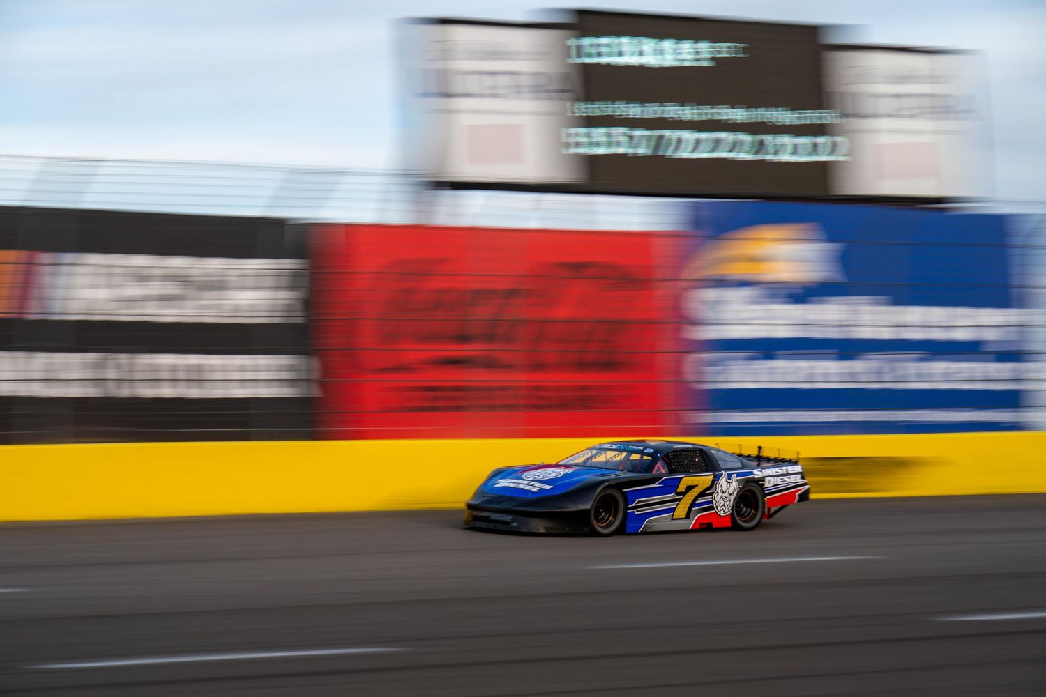 Blue and yellow NASCAR race car speeding past Coca-Cola advertisement boards on a professional oval racing track.