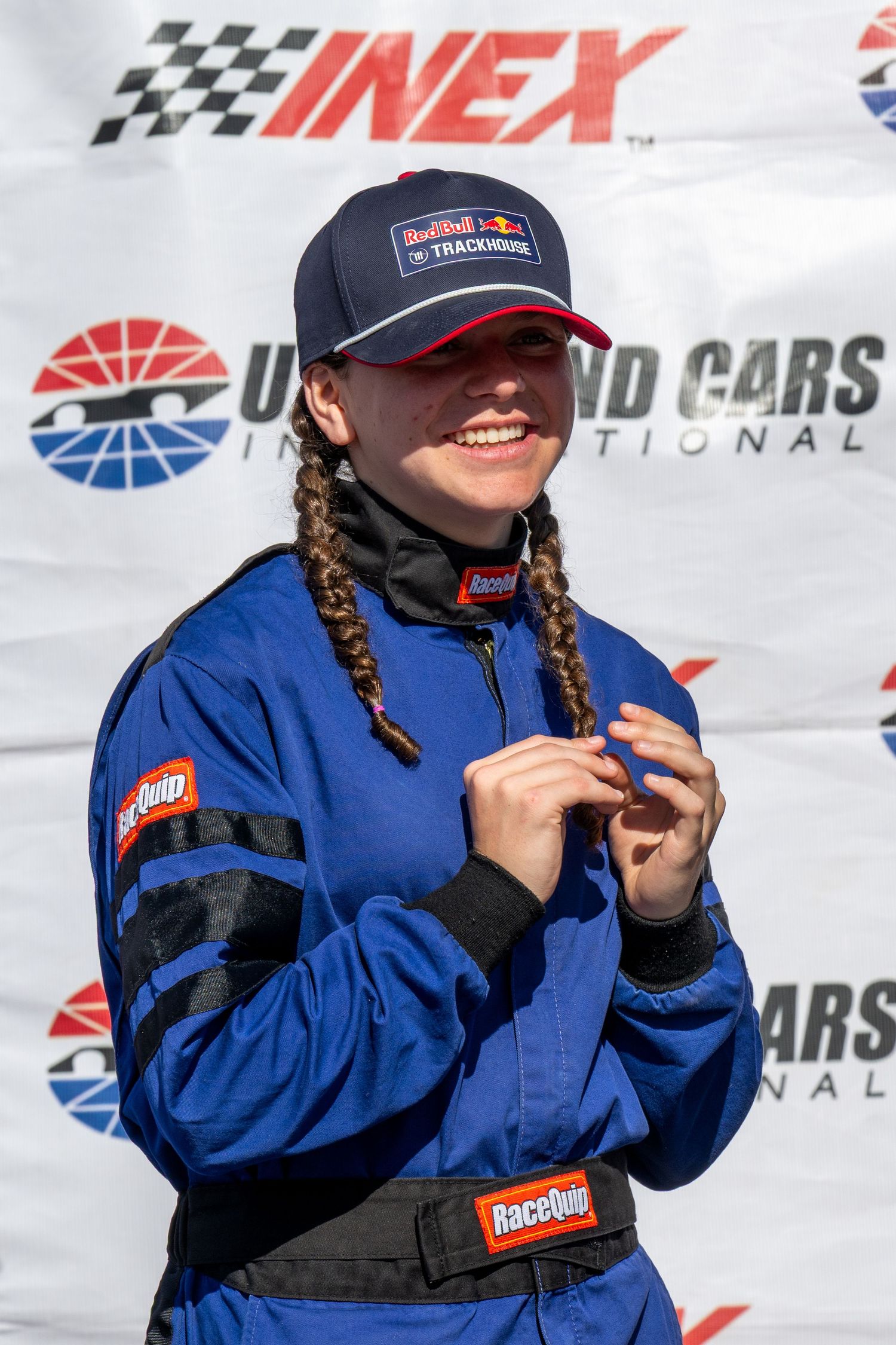 Female NASCAR driver in blue RaceQuip suit and Red Bull Trackhouse cap smiling at racing event.