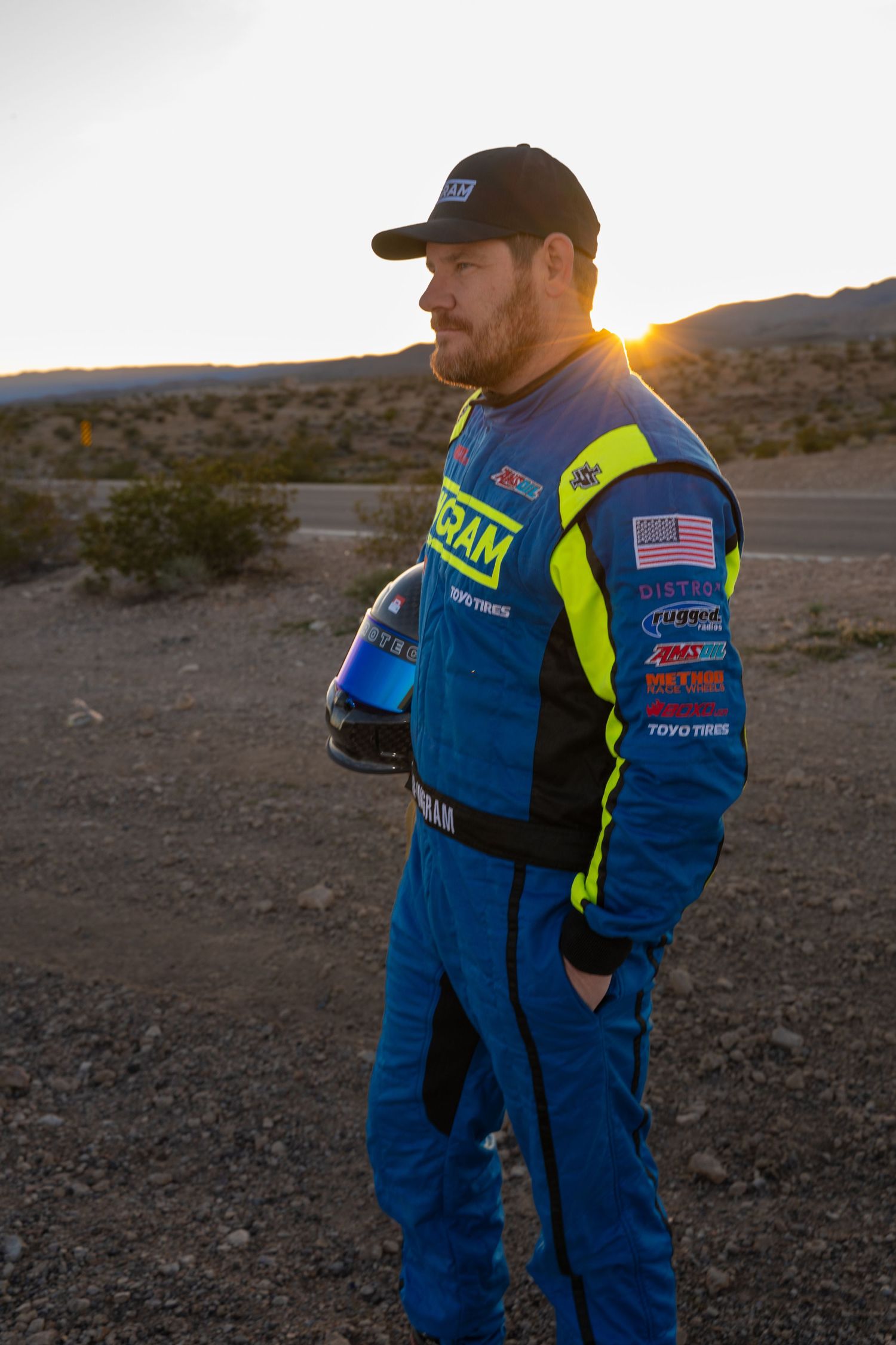 Off-road racing driver Chad Ingram in blue and yellow race suit holding helmet at sunset in desert landscape.
