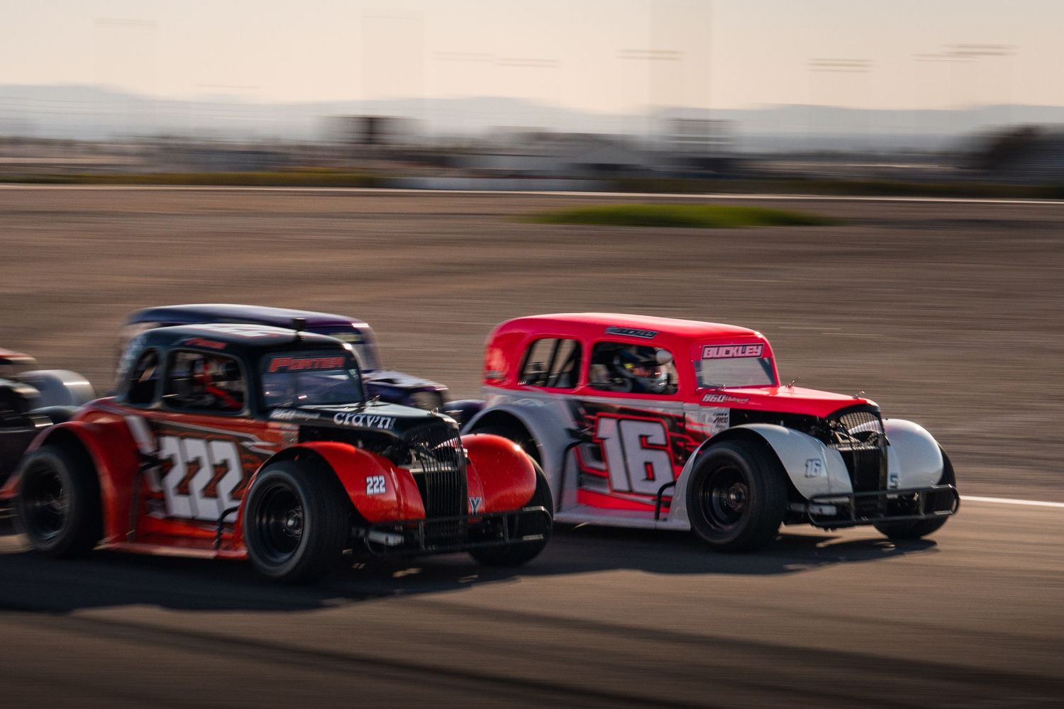 Two vintage legend cars, numbers 222 and 16, racing side by side on oval track.
