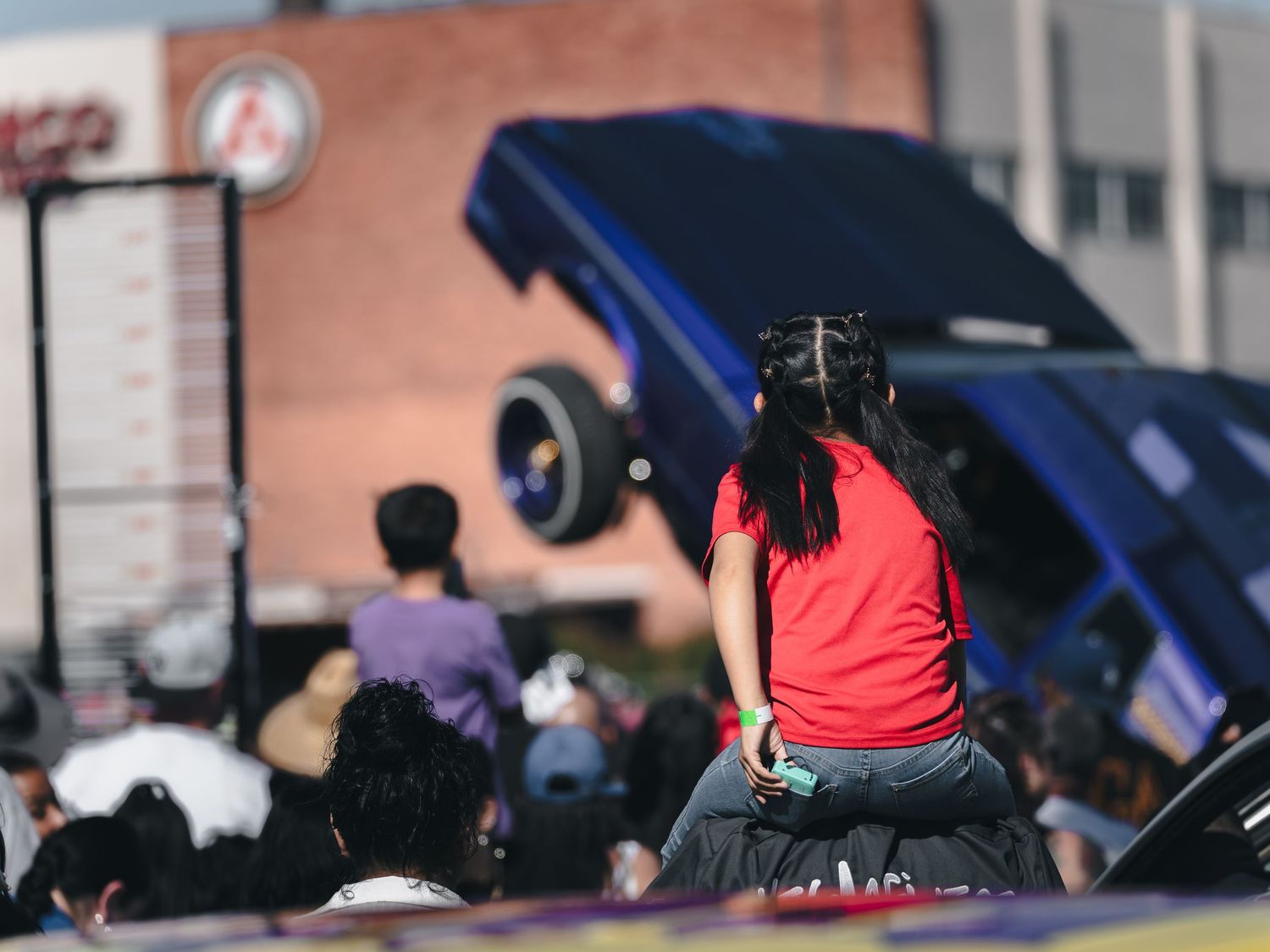Girl in red shirt sitting on lowrider car watching blue truck perform hydraulic hop at Super Saturday Weekend at The STRAT in Las Vegas