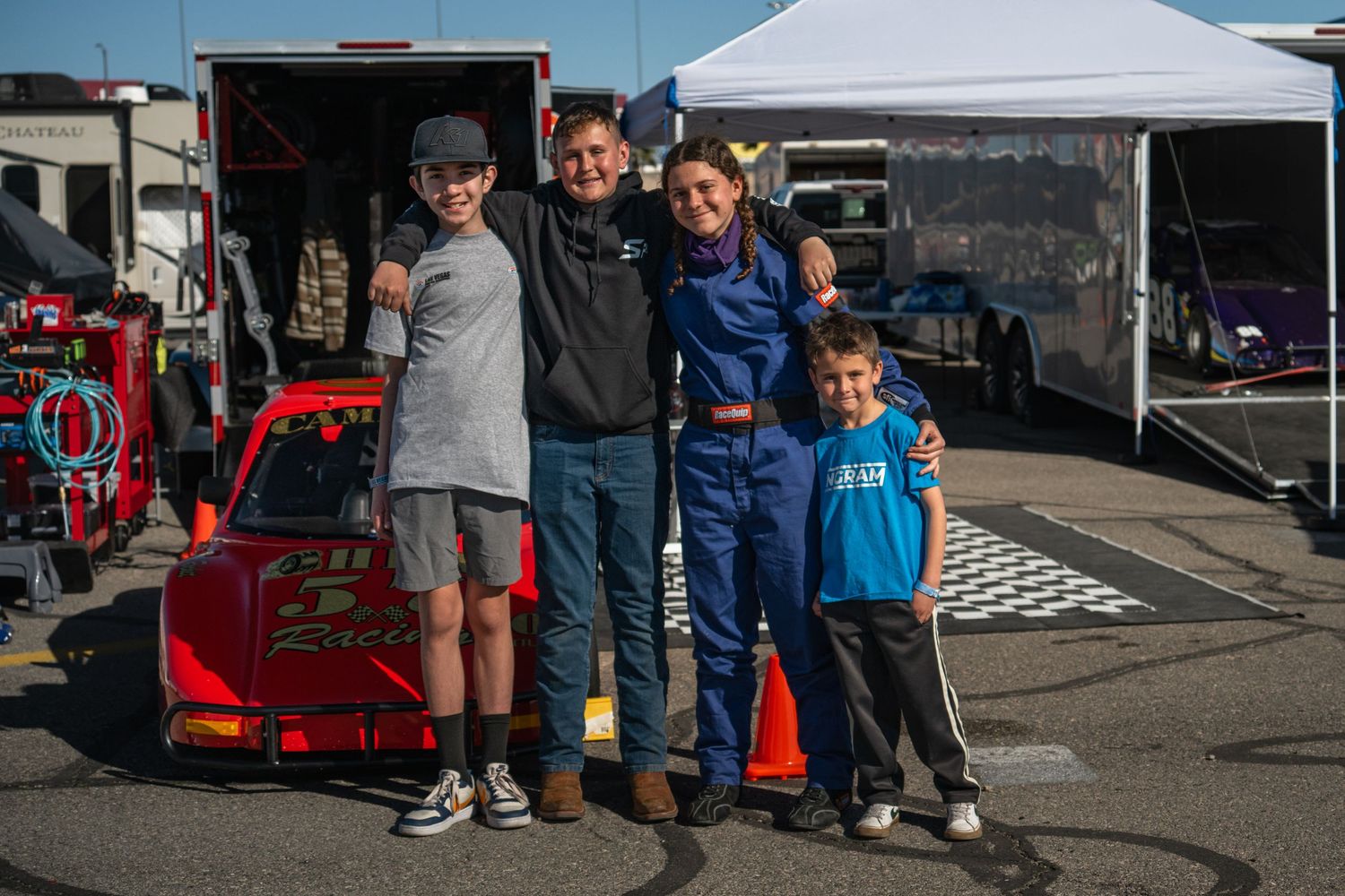 Family of four smiling together in a race track pit area beside a red race car.