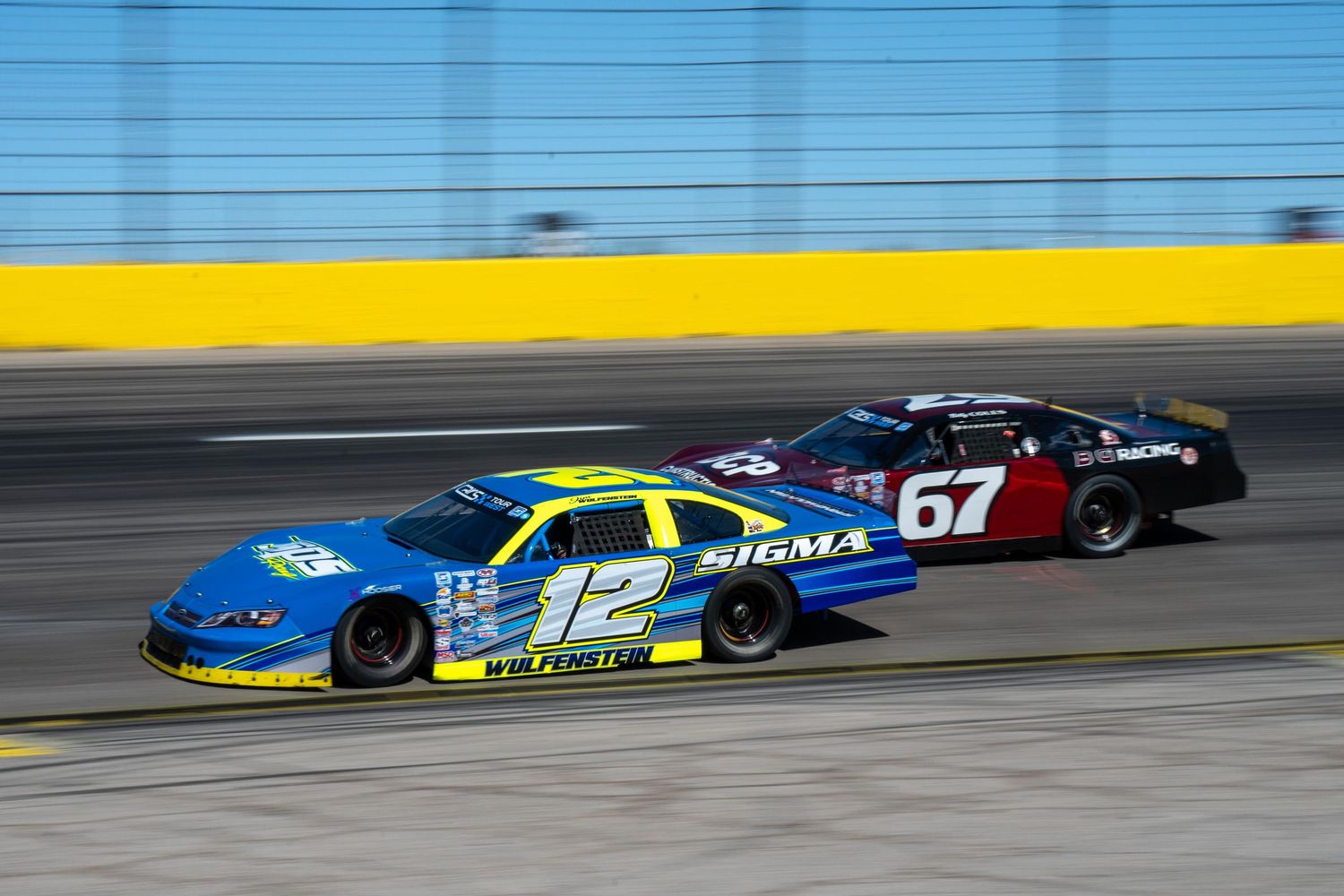 NASCAR stock cars number 12 and 67 racing side by side on a banked oval track during competition.