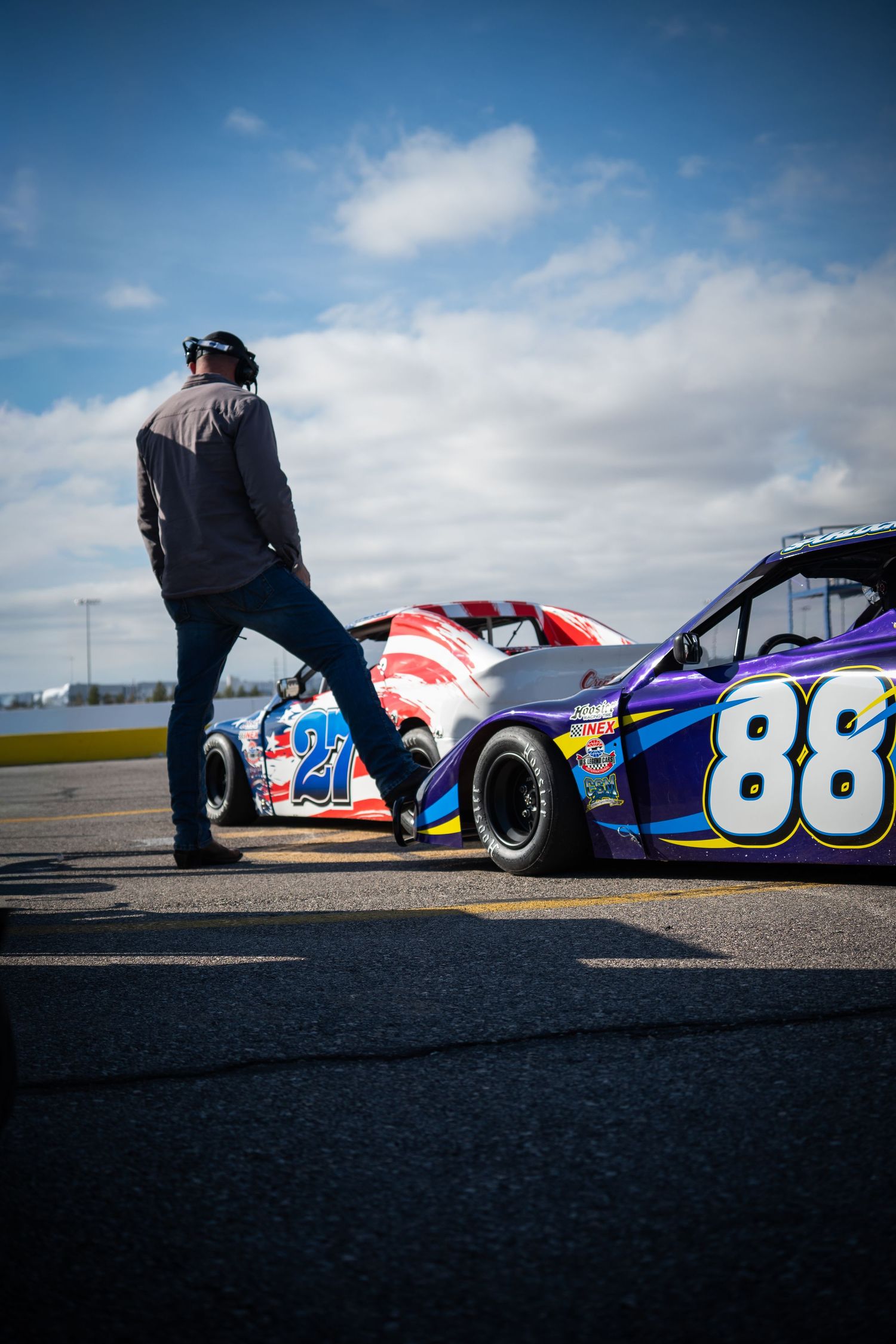 Man standing between two NASCAR race cars numbered 27 and 88 on a sunny racetrack pit lane.