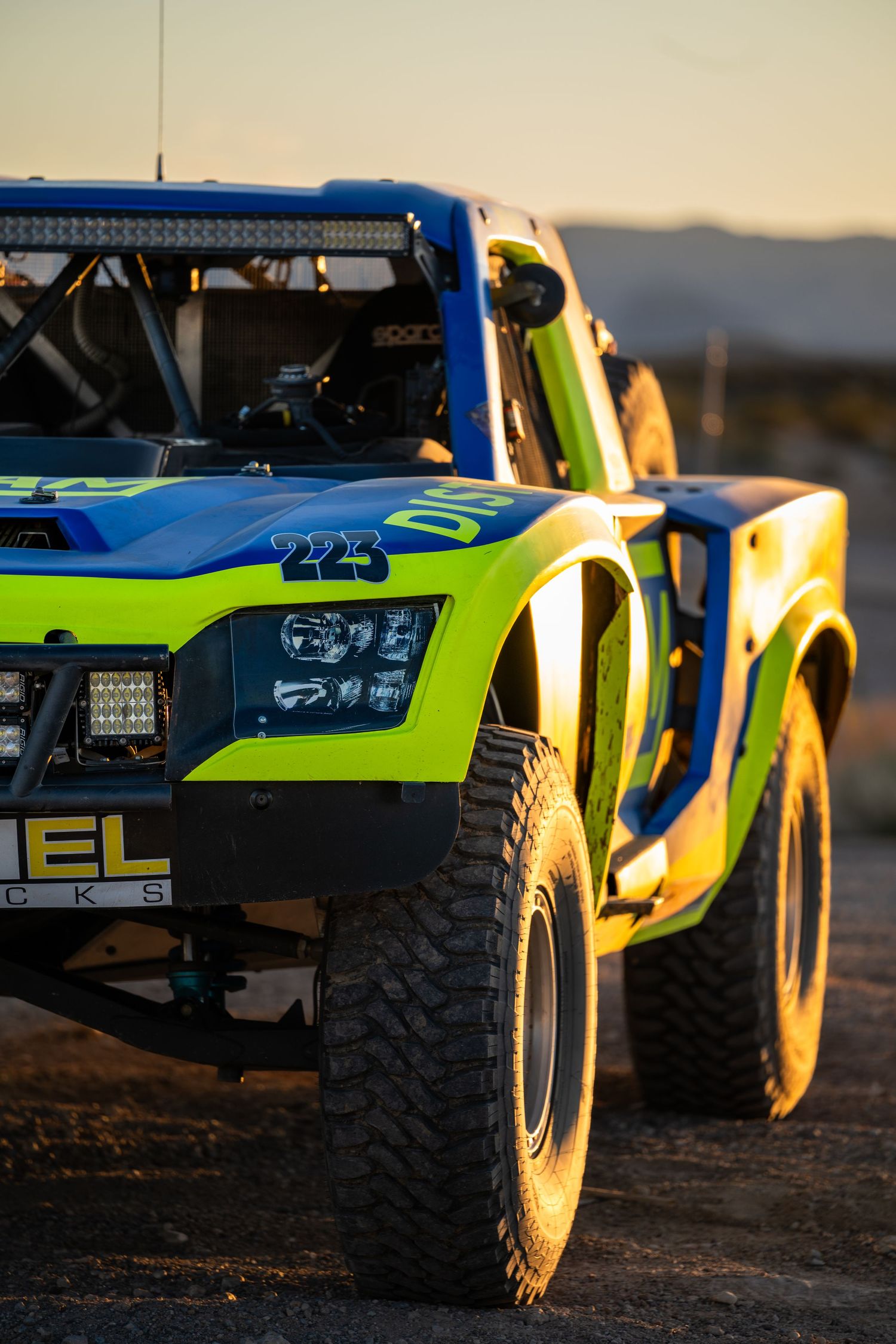 Front close-up of blue and green Ingram number 223 trophy truck at golden hour in desert setting.