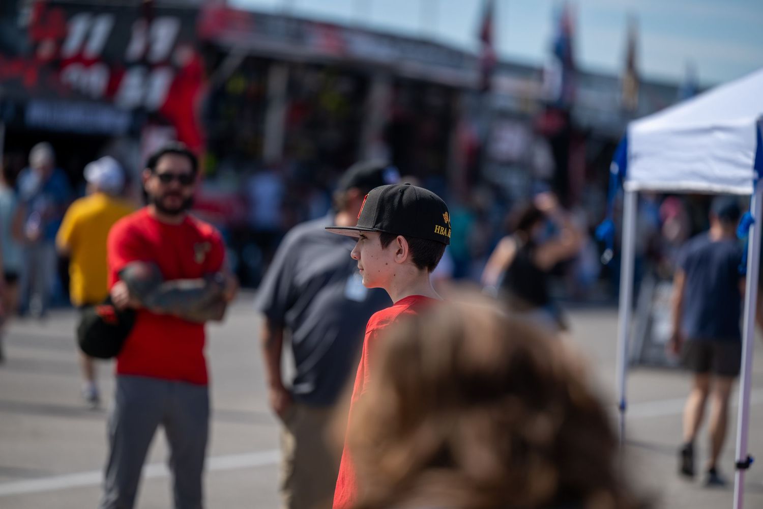 father and son in red shirt and cap at a NASCAR racing event with fans and vendor tents in background.