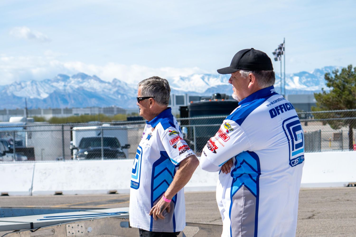 Two officials in blue and white racing uniforms stand trackside with snow-capped mountains in background.