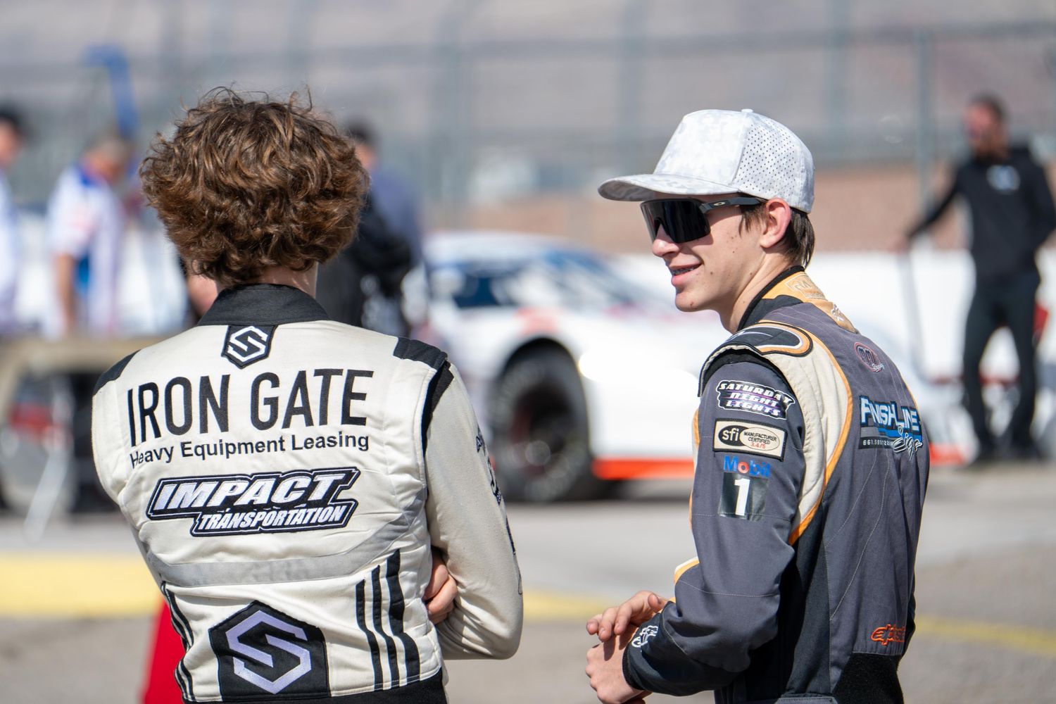 Two NASCAR drivers in racing gear share conversation on pit lane before race event.
