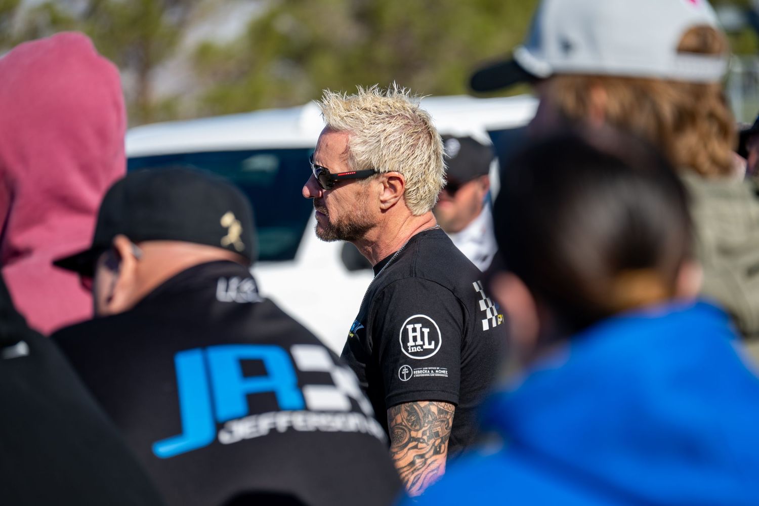 Tattooed man with blonde hair and sunglasses stands among crowd at outdoor motorsport racing event.