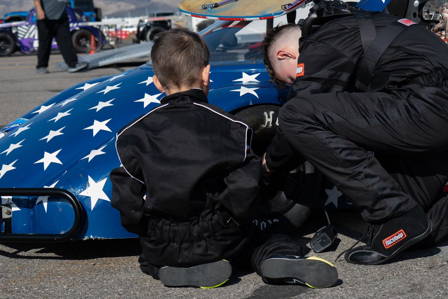 Adult and young child in racing gear crouching beside blue star-spangled race car on track.