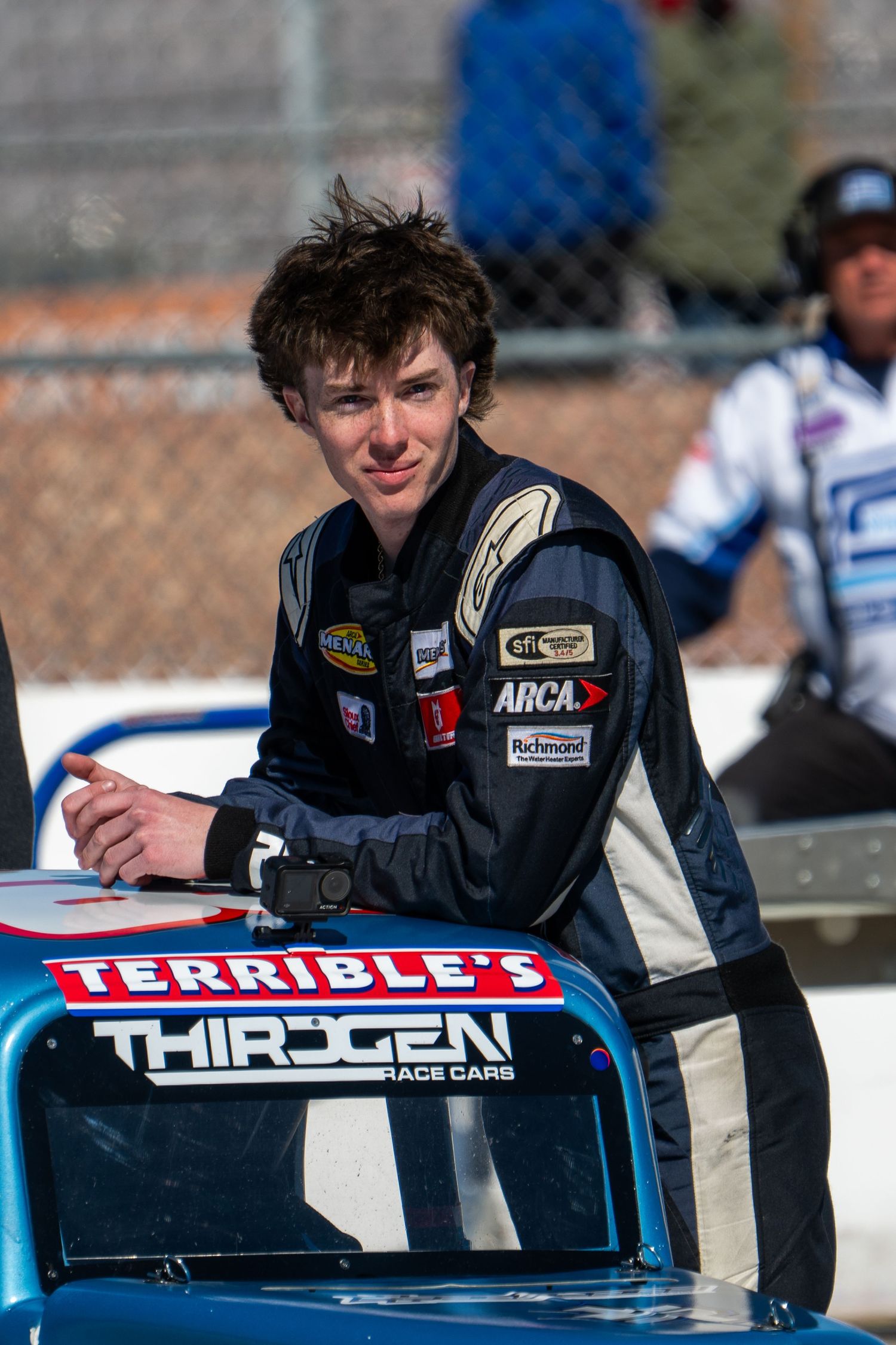 Young male race driver in race suit leans on Terrible's ThirdGen Race Cars vehicle.