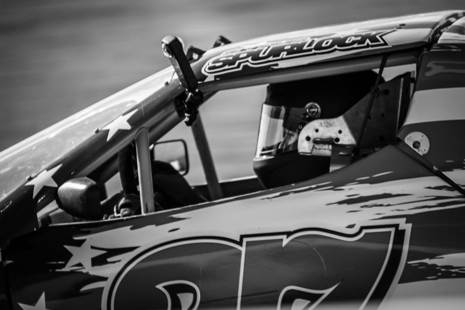 Black and white close-up of a NASCAR driver in helmet inside Spurlock race car cockpit.