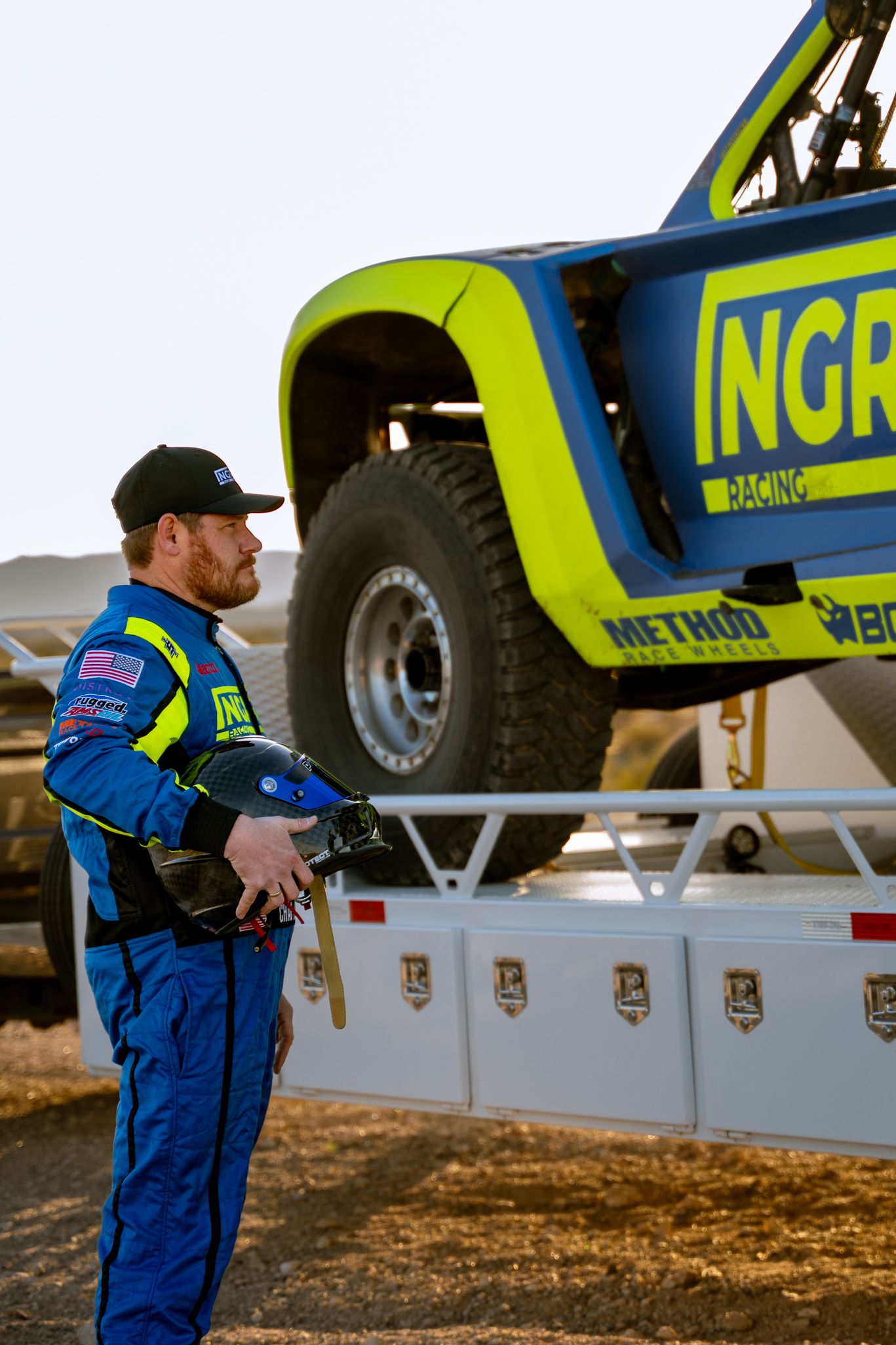 Off-road racing driver in blue suit watches Ingram Racing trophy truck being unloaded from trailer.