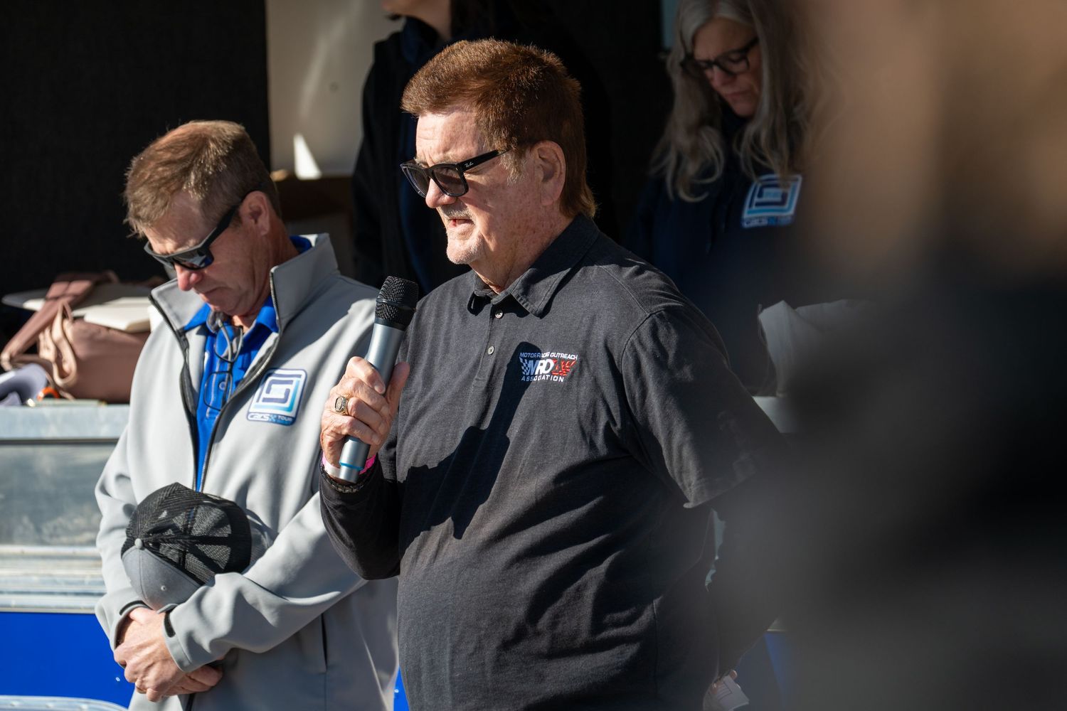 Man in dark racing jacket speaks at microphone during an outdoor motorsport event ceremony.