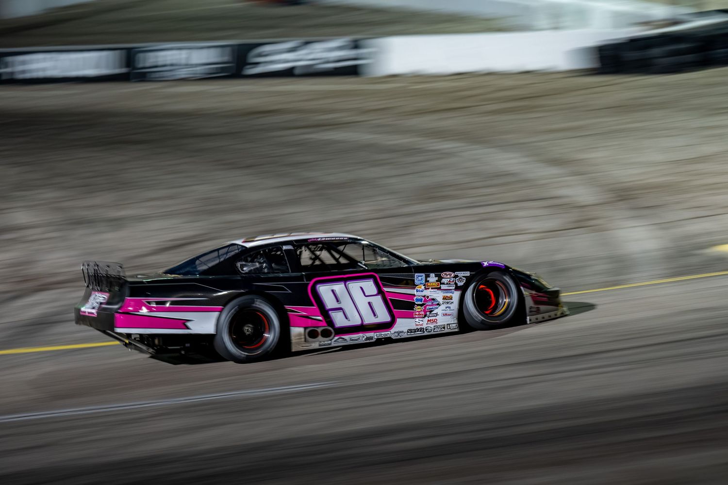 Black and pink number 98 prolate model race car speeding on dirt track during motorsport competition.