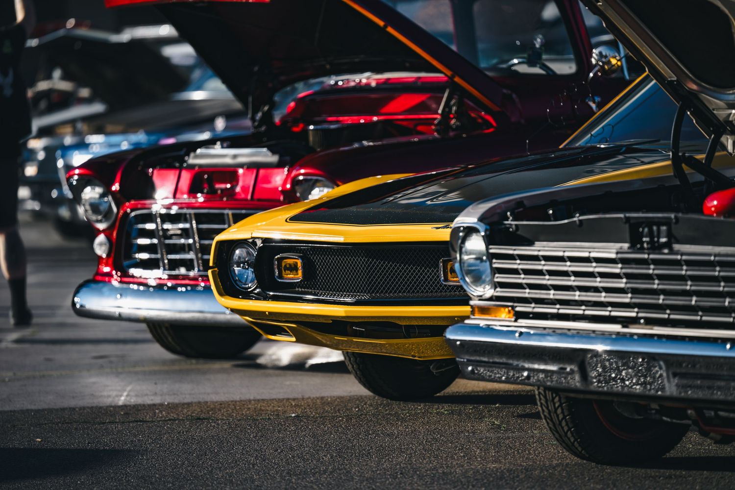 Classic cars with open hoods on display at a vintage car show, featuring red and yellow muscle cars.