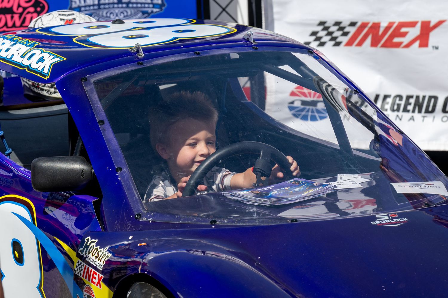 Adorable toddler sits behind the steering wheel of a purple bandolero at NASCAR