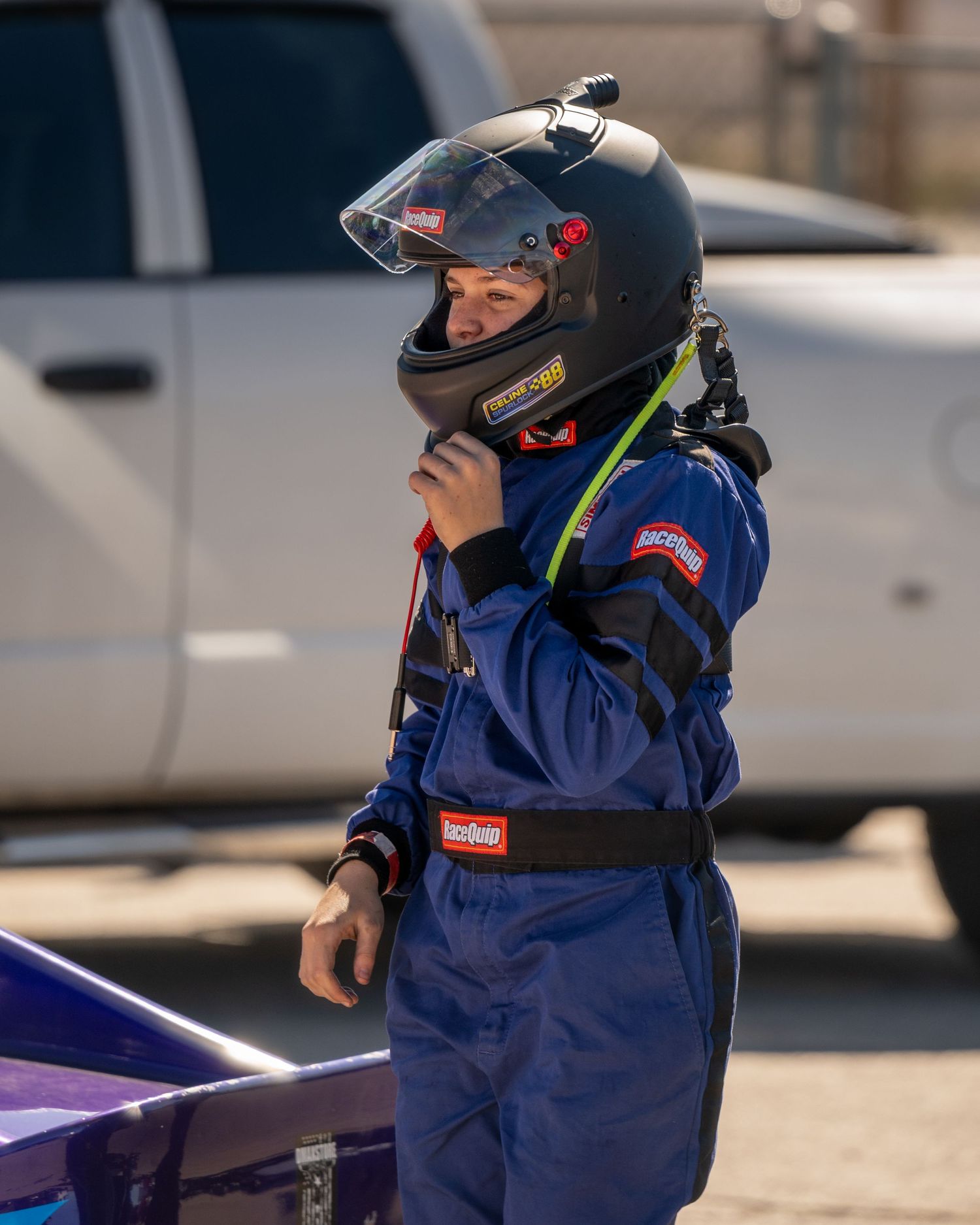 Young racing driver in blue RaceQuip fire suit adjusting helmet strap before a race event.