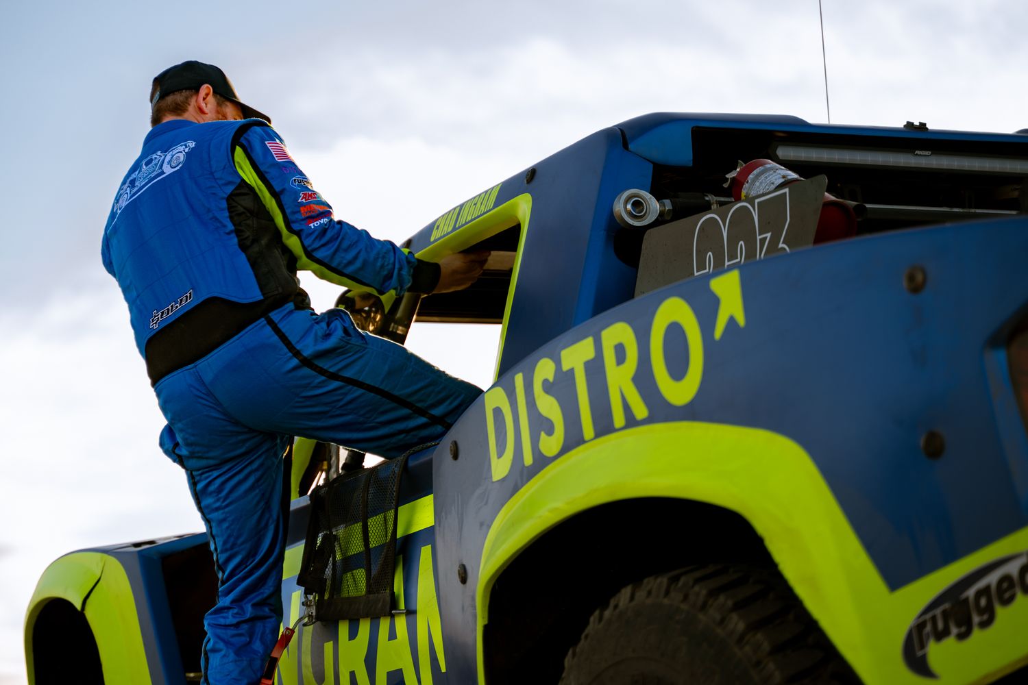 Racing driver in blue suit climbing into a Distro-sponsored off-road trophy truck.