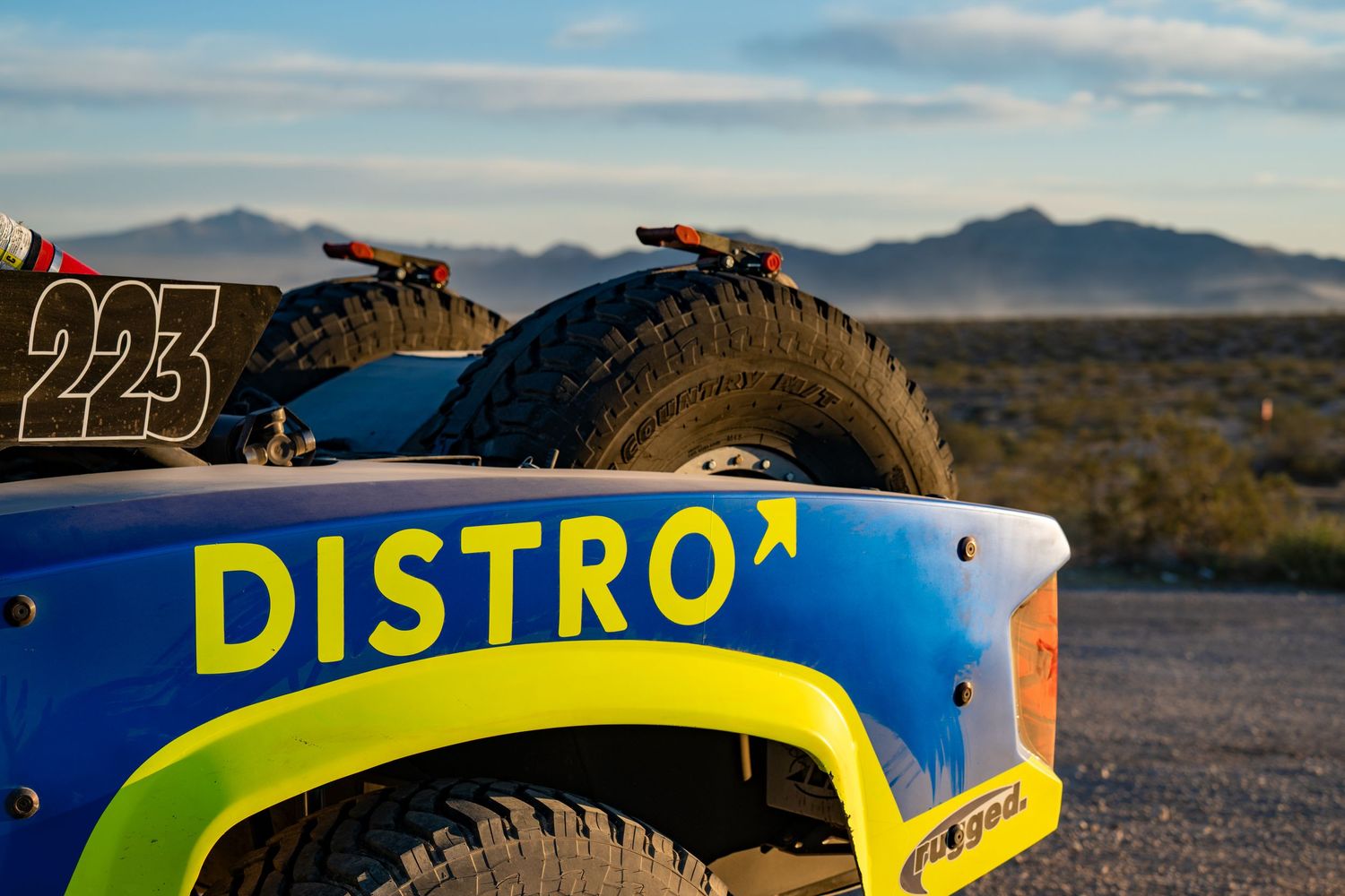 Rear view of blue and green Distro-sponsored trophy truck number 223 in desert landscape.