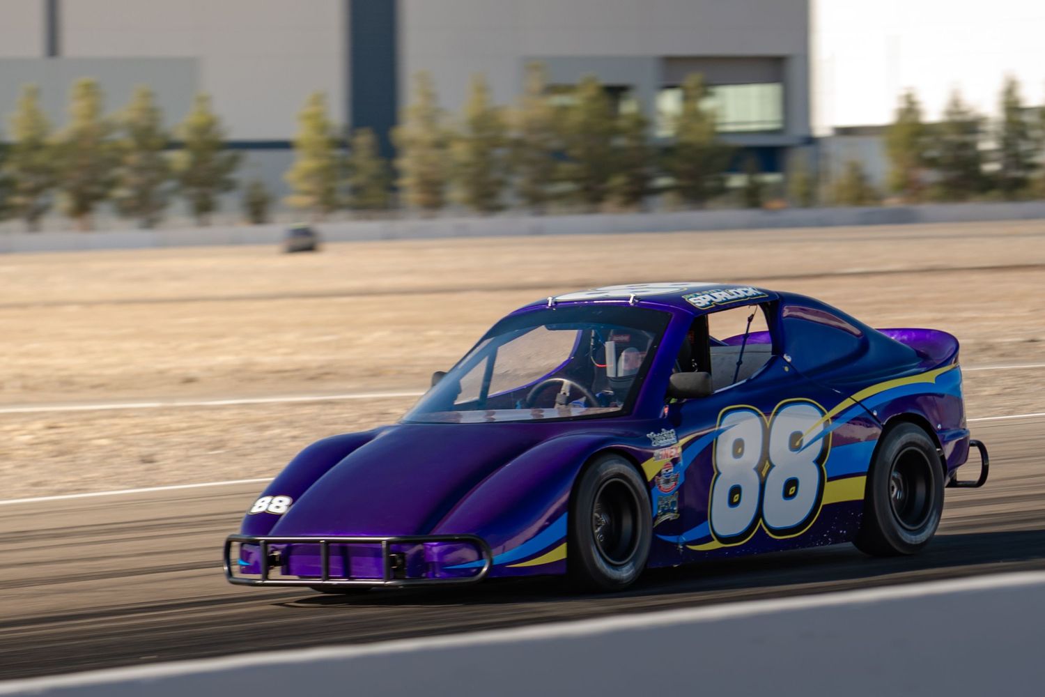 Purple and blue number 86 stock car racing on track at sunset during a motorsport event.