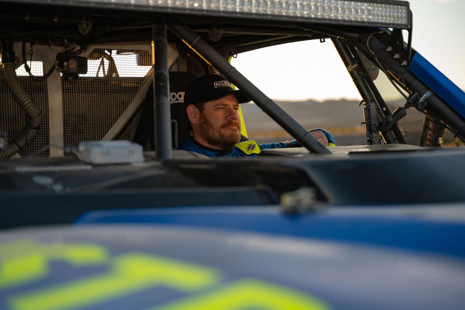 Male racing driver sitting inside a blue off-road trophy truck cockpit at sunset.